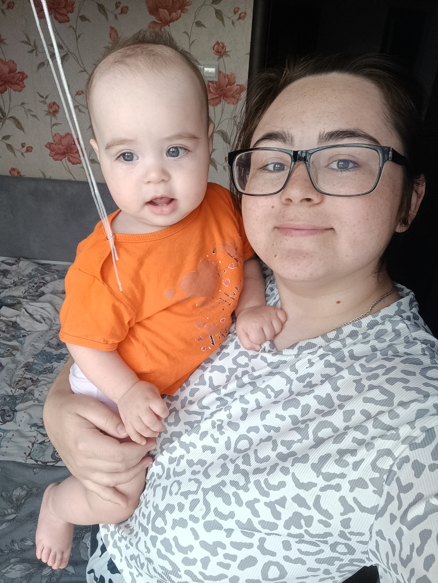A person wearing glasses and a grey and white leopard print shirt is holding a baby wearing an orange shirt. The baby has fair skin and blue eyes and is looking slightly to the left. The person's face is visible, and they are smiling slightly. The background shows a wall with floral wallpaper and a bed with patterned sheets. The image appears to be a selfie taken indoors. The city of Noviy, Russia is not visible in the image.
