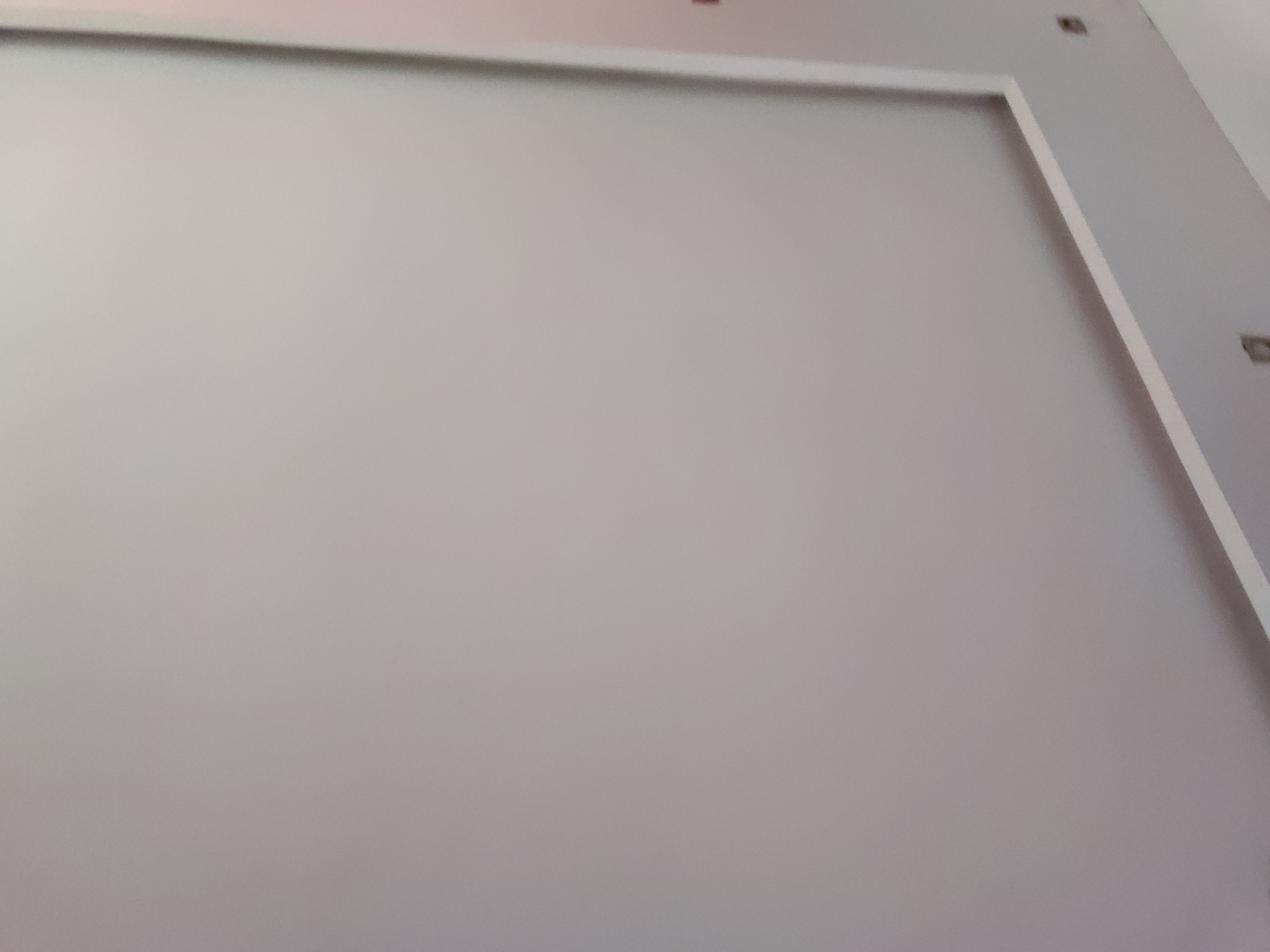 The image captures an interior wall, primarily in a uniform light grey hue. A prominent white, raised trim or molding outlines a rectangular section of the wall, extending horizontally across the top portion and then vertically downwards along the right side, creating a framed effect.
Several small, dark, square-shaped fixtures are subtly embedded into the wall. One is visible near the top edge, above the horizontal white trim. Two additional identical fixtures are positioned along the upper right edge of the wall, outside the vertical white trim. These fixtures appear to be recessed components, possibly lights or sensors.
The wall surface is smooth and unadorned, suggesting a modern or minimalist aesthetic. There are no people, objects other than the architectural elements, or discernible activity taking place. The lighting is even, and the scene is static. No text is visible in the image. While context suggests Santa Ana, Venezuela, there are no specific visual cues within the image itself to confirm this location. The overall impression is one of clean lines and neutral tones within an indoor setting.