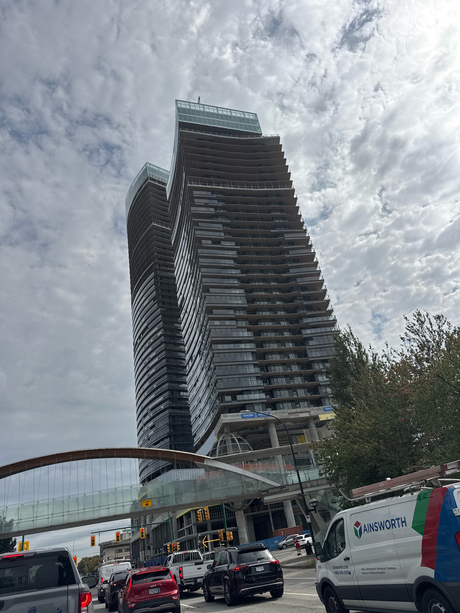 The image shows a view of two modern, high-rise buildings under construction in Burnaby, Canada, against a cloudy sky. The buildings have distinctive curved facades and appear to be residential or commercial towers. A skywalk or pedestrian bridge with a curved glass enclosure spans across the foreground, connecting to the buildings.
In the foreground, there is heavy traffic on a street. Several vehicles are visible, including a grey truck, a red SUV, a black SUV, a white van with "AINSWORTH" branding, and a white pickup truck. The Ainsworth van appears to be a service vehicle for HVAC, refrigeration, mechanical, plumbing, and electrical services, with contact information displayed on its side.
Traffic lights are visible, and street signs indicate an urban environment. Trees with green and some autumnal foliage are present on the right side of the image, partially obscuring the buildings. The overall scene suggests a busy urban setting with ongoing development.