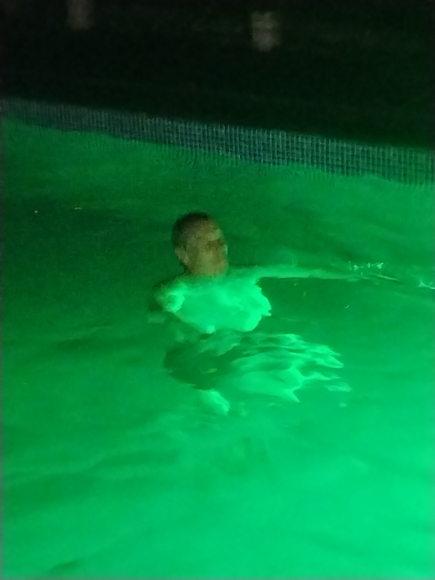 A person is swimming in a pool lit with green underwater lights. The person's head and upper body are visible above the water. The pool appears to be outdoors, with a dark, possibly natural, background behind it. The water is illuminated by the green lights, creating an eerie glow.
