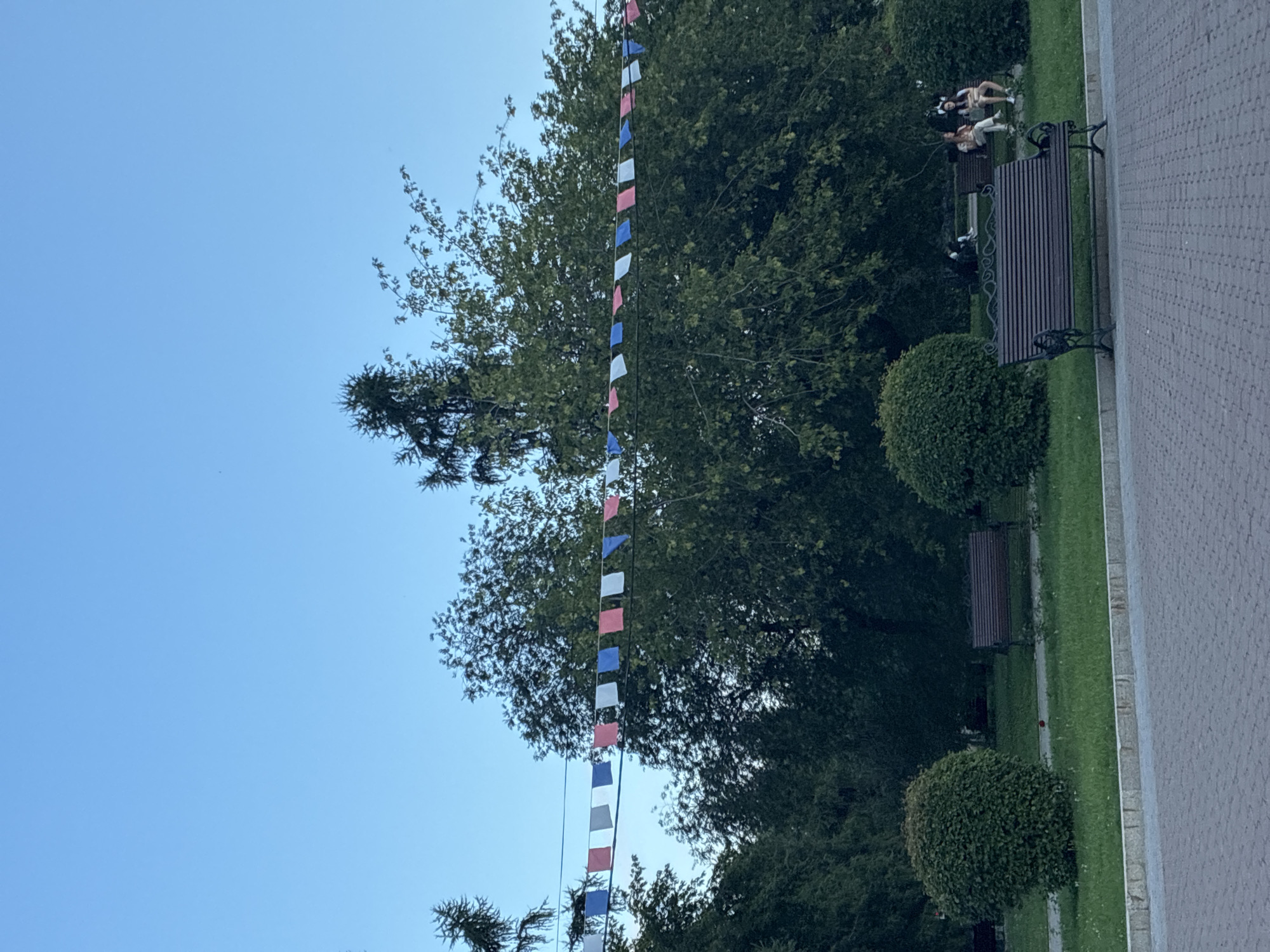 The image depicts a park scene in Irkutsk, Russia, under a clear blue sky. A string of colorful flags, in shades of red, white, and blue, stretches across the upper portion of the frame, strung between trees. Lush green trees and bushes dominate the midground, with several park benches visible. On one bench in the background, three individuals are seated, engaged in conversation or relaxation. In the foreground, a paved walkway is visible, suggesting a public park or recreational area. The overall atmosphere is calm and serene, with the natural elements complemented by the festive flags.