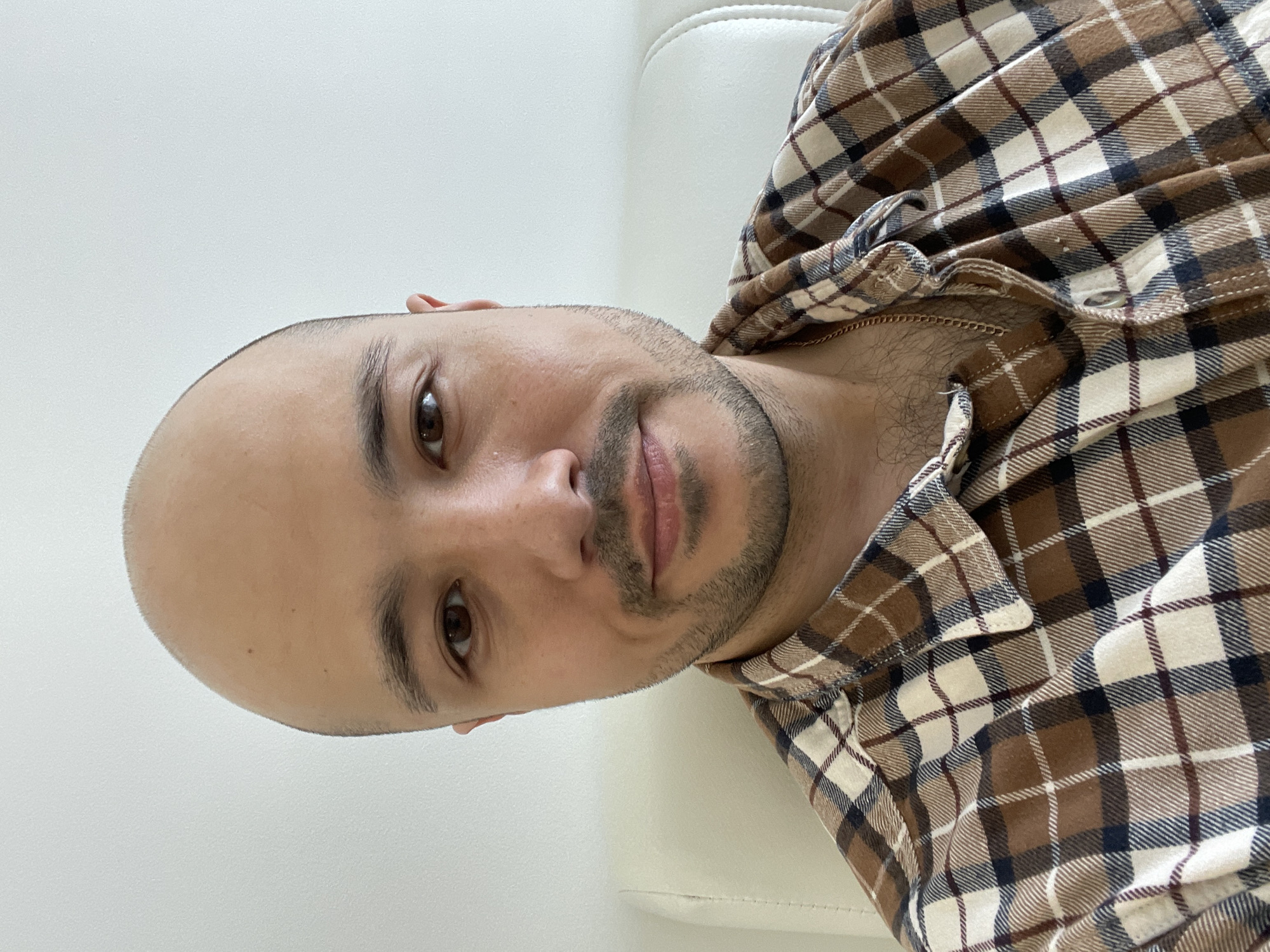 A close-up, low-angle head-and-shoulders shot depicts a man with a shaved head, dark eyebrows, brown eyes, a mustache, and a beard. He is wearing a buttoned plaid shirt, with brown, tan, and navy-blue squares. He is resting on a white surface, possibly a couch or cushion. The background is a plain white wall. The image does not provide sufficient information to determine the location as Izhevsk, Russia, or any other specific place.