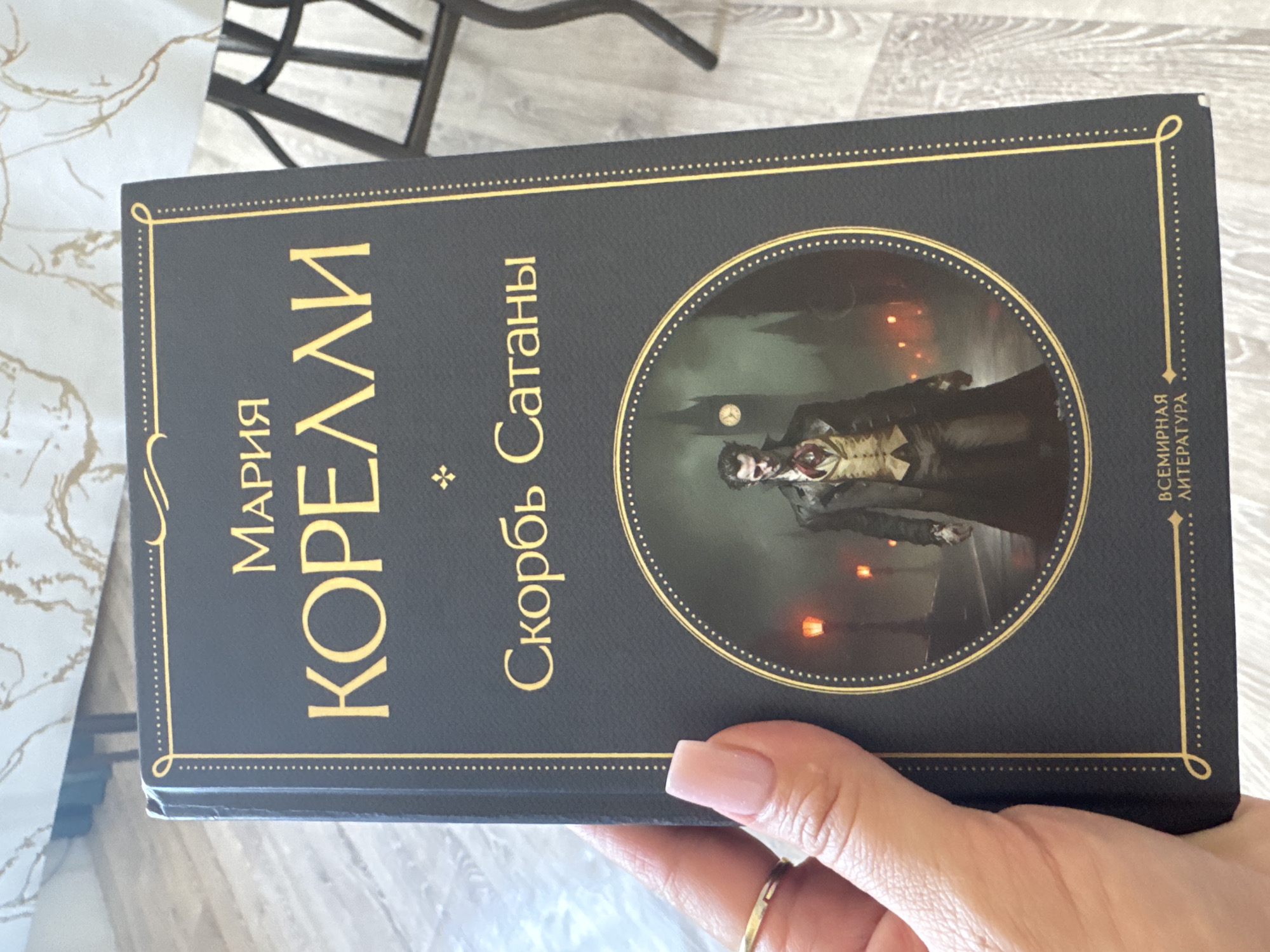 A dark blue book cover with gold lettering is held in a person's left hand. The cover features the title "Мария Корелли" above a circular illustration depicting a man in Victorian attire standing on a foggy street at night, with buildings and a clock tower in the background. Below the illustration, the text "Всемирная литература" is printed. The background of the image consists of a textured surface, possibly a table or floor, and the leg of a dark chair. There is no indication of the city of Jibek Joly, Kazakhstan, in the image.