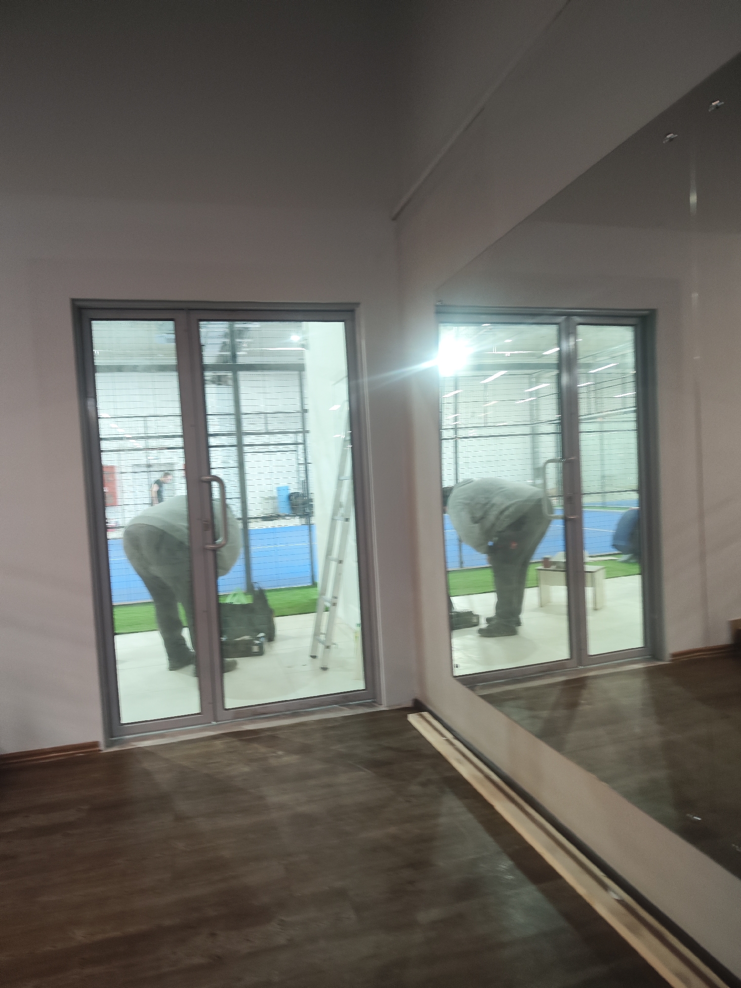 The image shows an interior view of what appears to be a commercial or sports facility in Chaikovsky, Russia. There are sliding glass doors that open into an area with a blue and green playing surface, enclosed by a wire mesh. A person in grey clothing is bent over, seemingly working on something inside the playing area. There is a stepladder next to them. The image also shows a reflection in a large mirror in the foreground, mirroring the view through the doors. The floor in the foreground has a dark, wood-like appearance.
