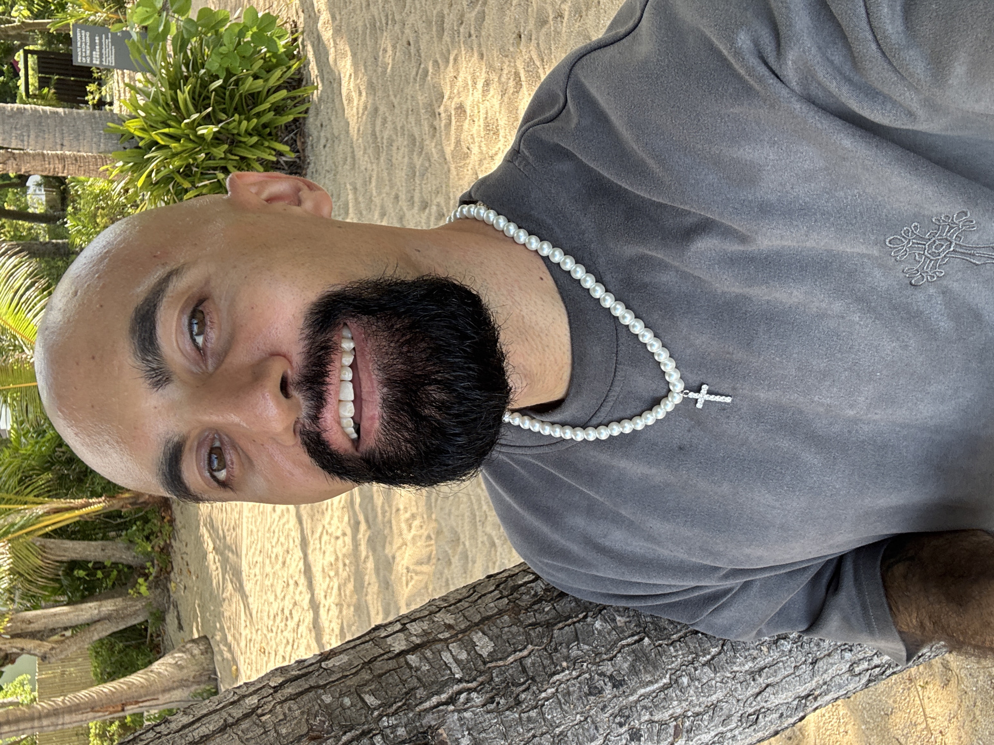 A bald man with a goatee and a pearl necklace with a cross pendant is standing on a sandy beach. He is wearing a gray t-shirt. Palm trees and lush green vegetation are visible in the background, along with a wooden fence and a sign. The sign in the background is in Thai and English and contains information about private property and no trespassing. The location appears to be a tropical beach in Koh Samui District, Thailand.