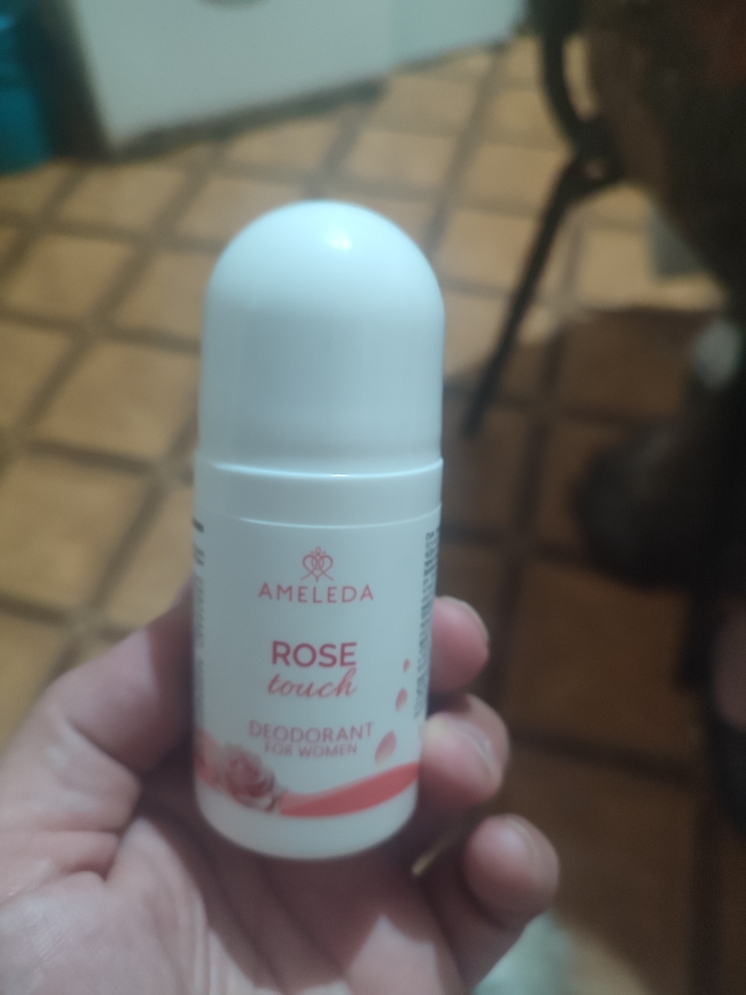 A close-up, eye-level shot shows a white roll-on deodorant bottle being held by a person's hand. The bottle has pink lettering and a stylized rose graphic. The brand name "AMELEDA" is visible above the text "ROSE touch," which is followed by "DEODORANT FOR WOMEN." The background is blurred, showing a tiled floor and indistinct furniture. The image appears to be taken indoors, possibly in a residential setting. There is no discernible indication of the location being Chernivtsi, Ukraine.