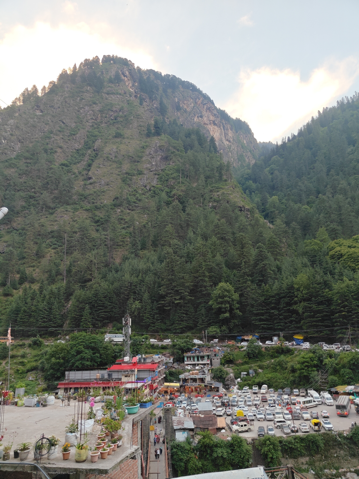 This vertical photograph presents a vibrant mountain townscape, characteristic of Manikaran, India, set within a verdant valley. Towering, rugged mountains, densely covered with coniferous trees, dominate the background, their rocky slopes rising steeply towards a cloudy sky.
In the mid-ground, a bustling settlement unfolds across the valley floor. A multitude of buildings with varied architecture can be seen, some featuring distinctive red roofs. A tall communication tower stands prominently near a larger building. The area below these structures is a large, crowded parking lot, packed with numerous vehicles including cars, vans, and buses, indicating a busy hub or tourist destination. People are observed moving about among the vehicles and on a pedestrian walkway or bridge to the right, suggesting active daily life. Faintly visible on a shop sign in the parking lot area are the words "Shree-Rajesh."
The immediate foreground features a rooftop adorned with a diverse collection of potted plants, some blooming with flowers like orchids, adding a touch of greenery to the built environment. Utility wires and cables stretch across the scene, connecting different parts of the settlement and traversing the mountain slopes.
The time of day appears to be either early morning or late afternoon, characterized by soft, diffused light filtering through the overcast or partly cloudy sky, casting no harsh shadows. The overall scene depicts a lively town integrated closely with its dramatic natural mountain surroundings.
