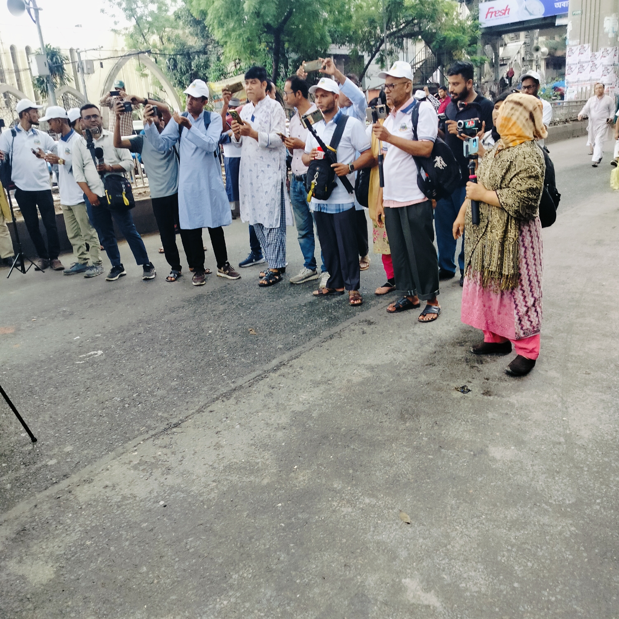 A medium shot shows a group of people, mainly men, standing on a paved street. They are mostly fair-skinned with dark hair, and many are wearing white baseball caps. Most are holding up cell phones or cameras, seemingly capturing an event. A woman in the foreground, draped in a patterned shawl and pink skirt, also holds a cell phone on a selfie stick. The group appears to be focused on something out of frame.
The scene is set against an urban backdrop with buildings and trees visible in the distance. An overhead sign reading "Fresh" is partially visible. In the background, an older building can be seen with architectural details such as archways. The presence of a tripod suggests a more formal recording set up. There is a sidewalk and possibly a crosswalk in the background. The location is in the city of Unknown, Unknown.