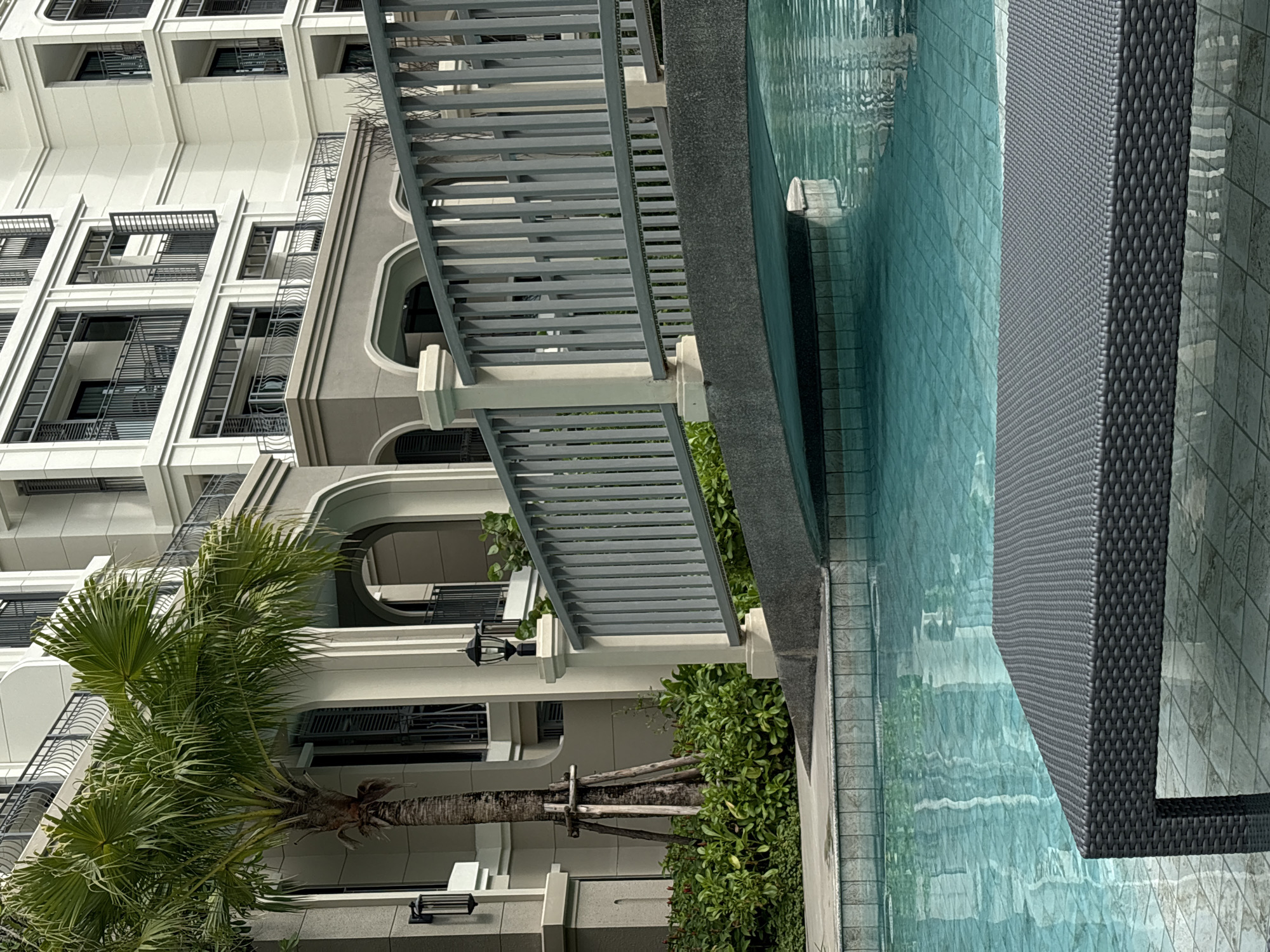 The image shows a luxury apartment building with a swimming pool in Choeng Thale, Thailand. The building is a multi-story structure with modern architectural features, including arched windows and balconies. A tall palm tree grows near the building, adding a tropical element to the scene. The swimming pool is adjacent to a paved area with a lounge chair. The water in the pool is clear and reflects the sky and surrounding structures.