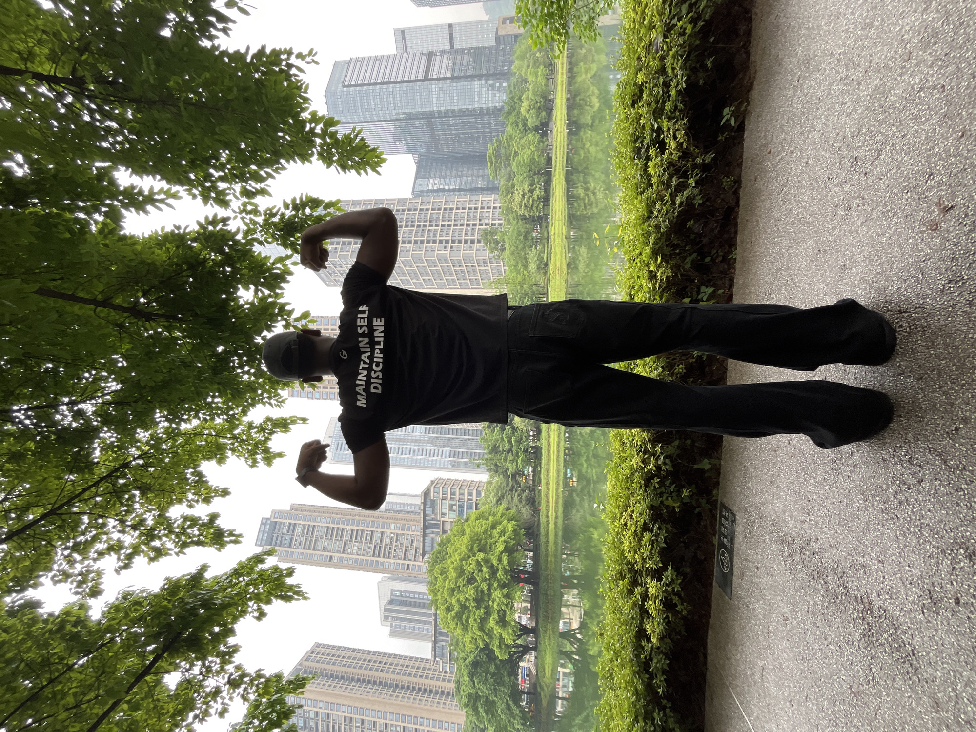 A man stands on a paved path, back to the camera, flexing his biceps. He wears a black t-shirt with the words "MAINTAIN SELF DISCIPLINE" printed on the back, black pants, and a baseball cap. In the background, modern high-rise buildings dominate the skyline of Yuzhong District, China, reflected in a body of water. Green trees line the periphery of the body of water, bordering the path. The weather appears overcast, with light filtering through the tree branches overhead.