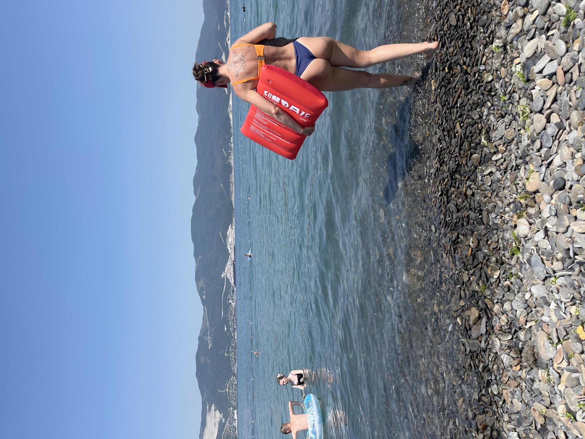 A woman in a blue bikini and an orange bikini top walks into the sea carrying a red inflatable float. She has dark hair pulled up and a red bandana. Mountains are visible in the background. Several people are swimming in the sea, including a child in an inflatable ring. The foreground is a pebble beach.