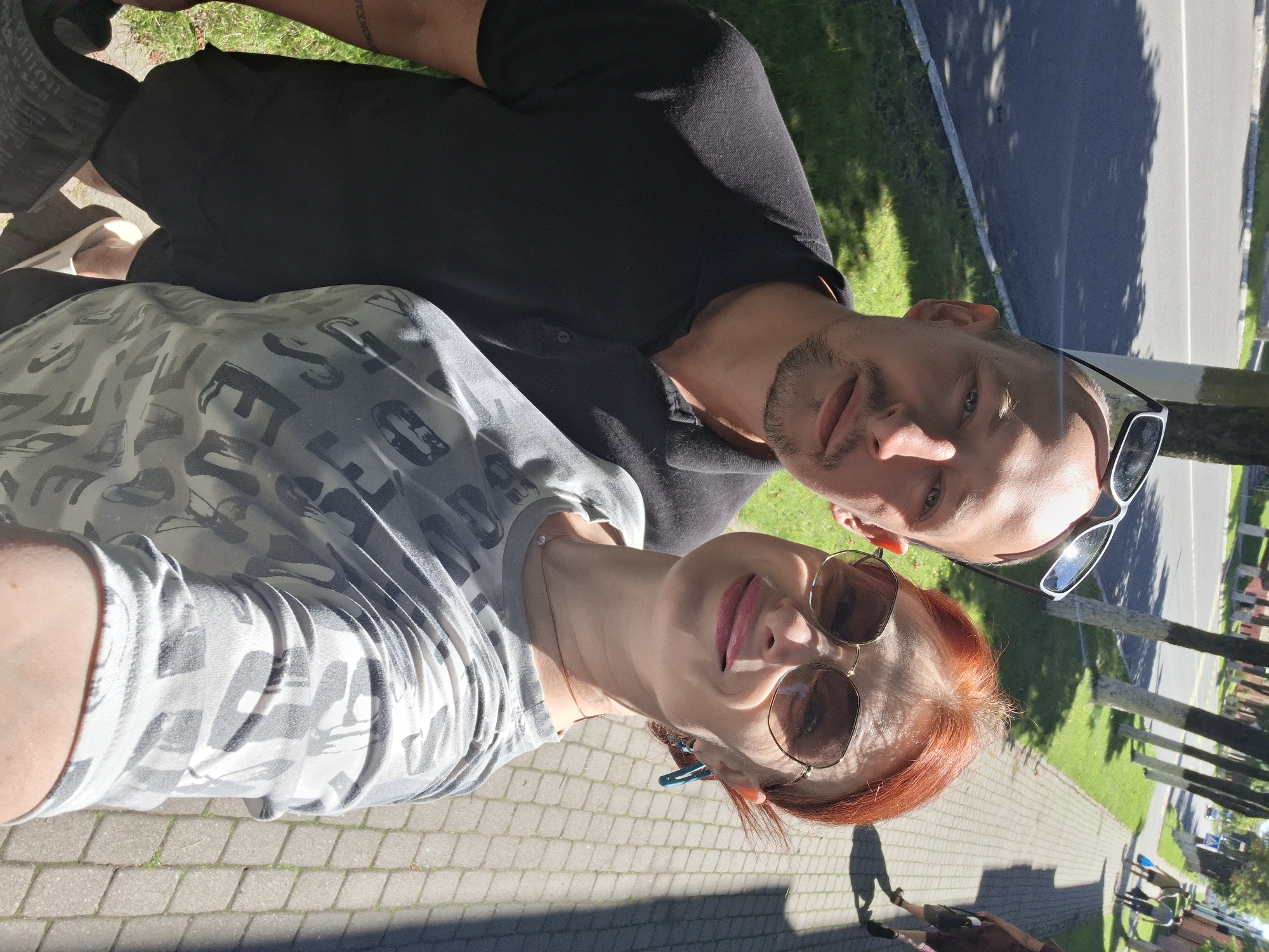This is a close-up, outdoor, selfie-style photograph of a couple standing on a paved walkway next to a street. The man, on the left, is wearing a black polo shirt and sunglasses perched on his head. He has a shaved head and a goatee. The woman, on the right, has red hair and is wearing large, round sunglasses and a grey t-shirt with a white graphic print. She is also wearing a small pendant necklace and large, teal-colored earrings. The background shows a tree-lined street with a sidewalk, green grass, and parked cars in the distance. The overall lighting suggests it is a sunny day. The scene is consistent with a casual daytime outing in the city of Myadzyel, Belarus.