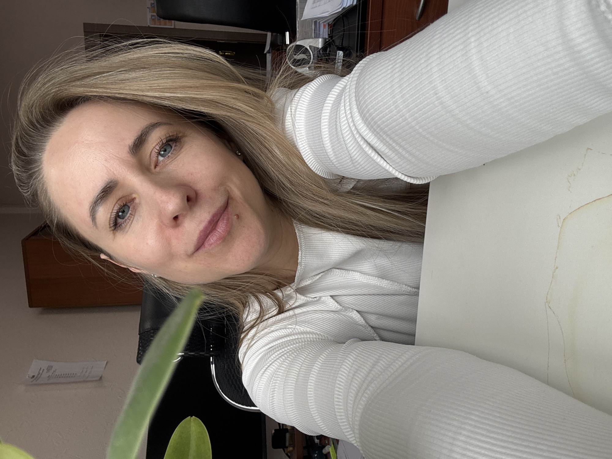 A blonde woman with fair skin and blue eyes is facing the camera. She has a mole on her left cheek. She is wearing a white, ribbed sweater. Her hair is parted to the side. A green plant is partially visible in the foreground. Behind her, there is an office setting with a black chair, dark wooden furniture, and documents on the wall. The image may have been taken indoors in the city of Chaikovsky, Russia.