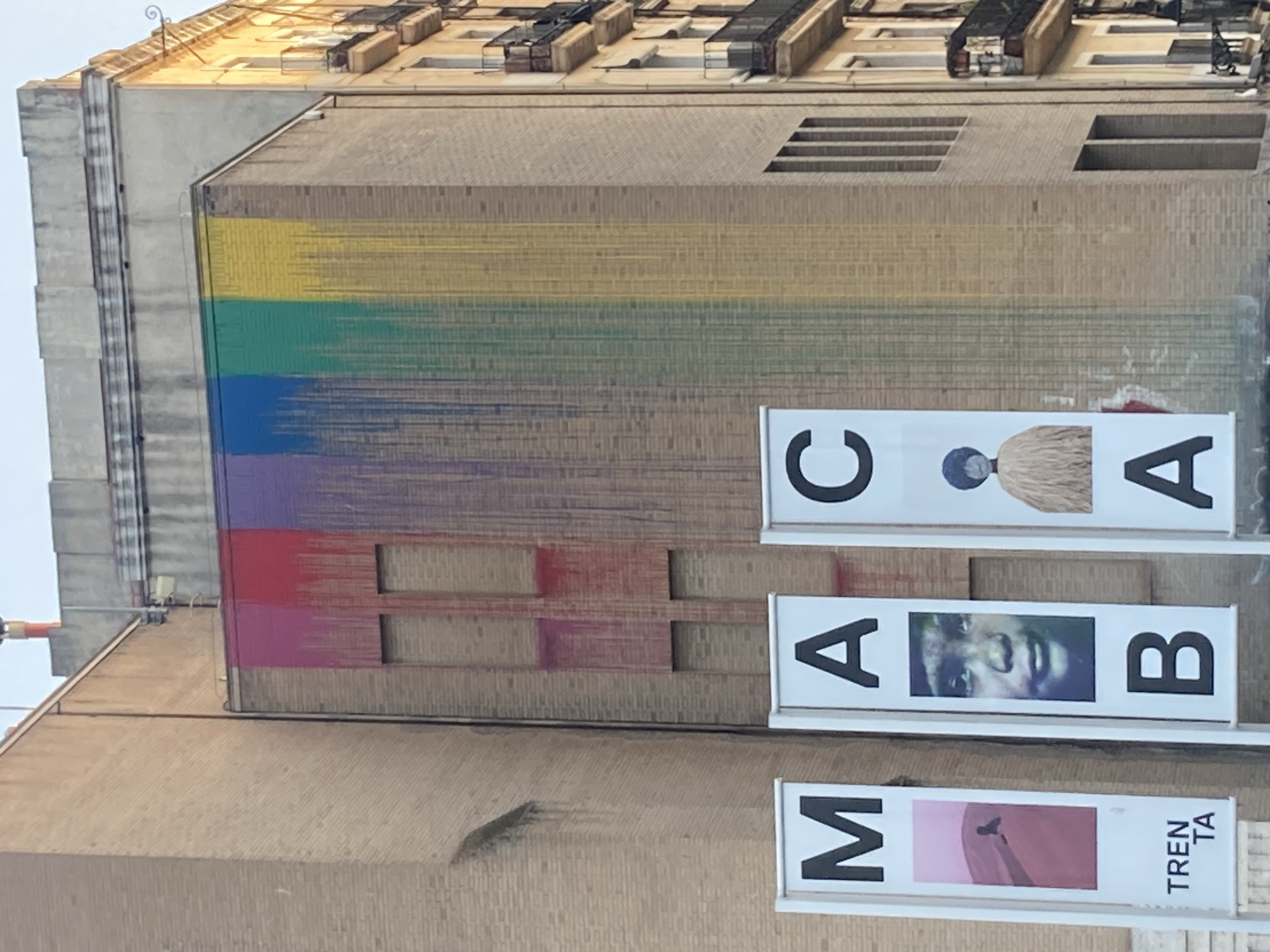 This image depicts the MACBA (Museum of Contemporary Art of Barcelona) building, located in the El Raval neighborhood of Barcelona, Spain. The museum's facade is adorned with a large, colorful mural that resembles a rainbow, with streaks of red, pink, purple, blue, green, and yellow paint dripping down the brickwork. In front of the building, three vertical banners are displayed. The left banner features a pink graphic with a silhouette of a figure and the text "TRENTA TA". The middle banner shows a black and white portrait of a child and the letters "M A B". The right banner displays a figure with a dark blue head and a shaggy, light-colored garment, accompanied by the letters "C A". The scene is illuminated by natural light, suggesting it was taken during the daytime.