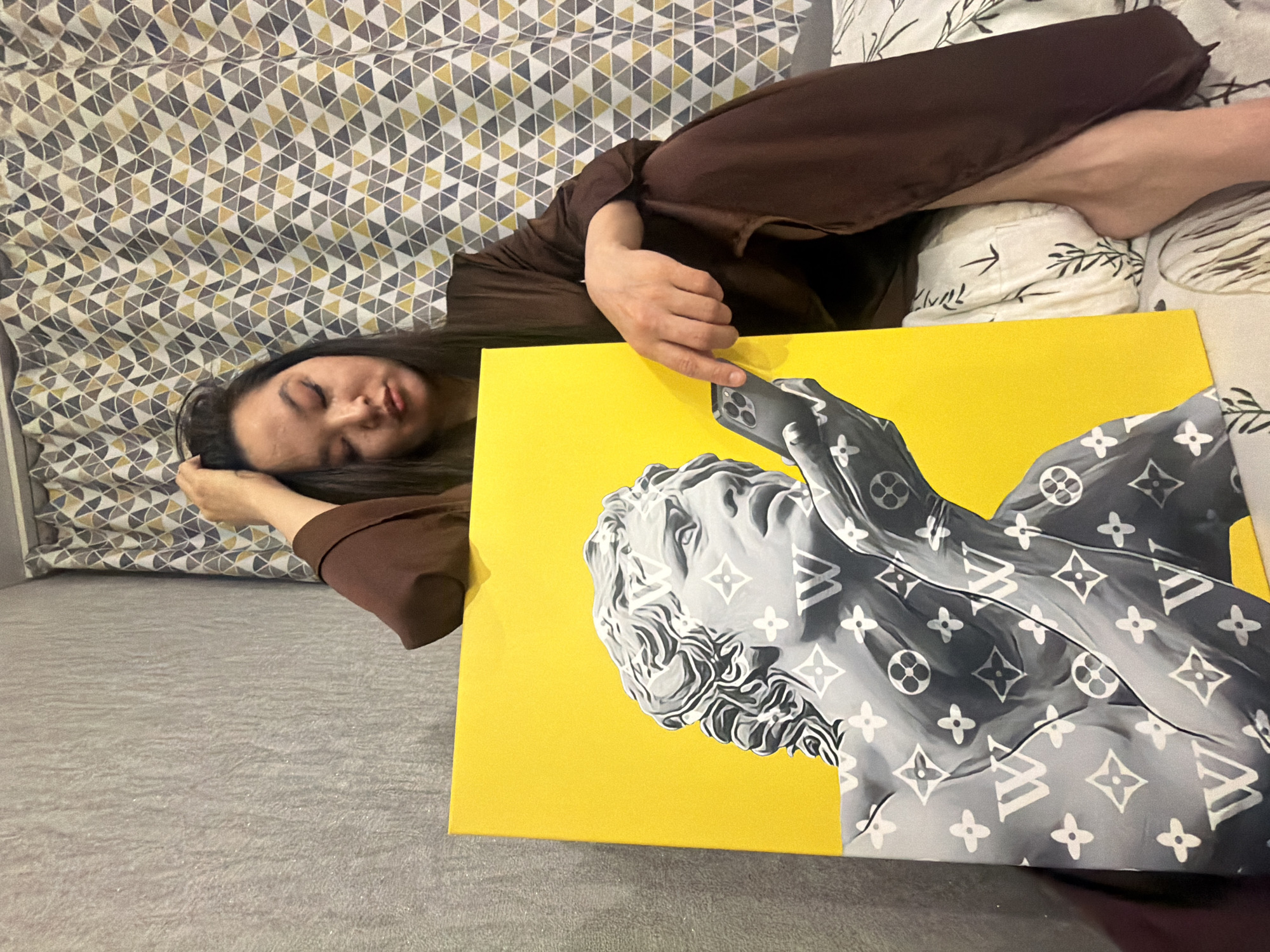 A person with long dark hair is lying down on a bed, with their head resting on a pillow with a geometric pattern. They are wearing a brown outfit. Next to them is a canvas painting with a yellow background, featuring a grayscale image of a classical bust design with a Louis Vuitton monogram pattern. The bed is covered with a grey textured fabric. The location appears to be inside a bedroom, but the specific city and location are unknown.