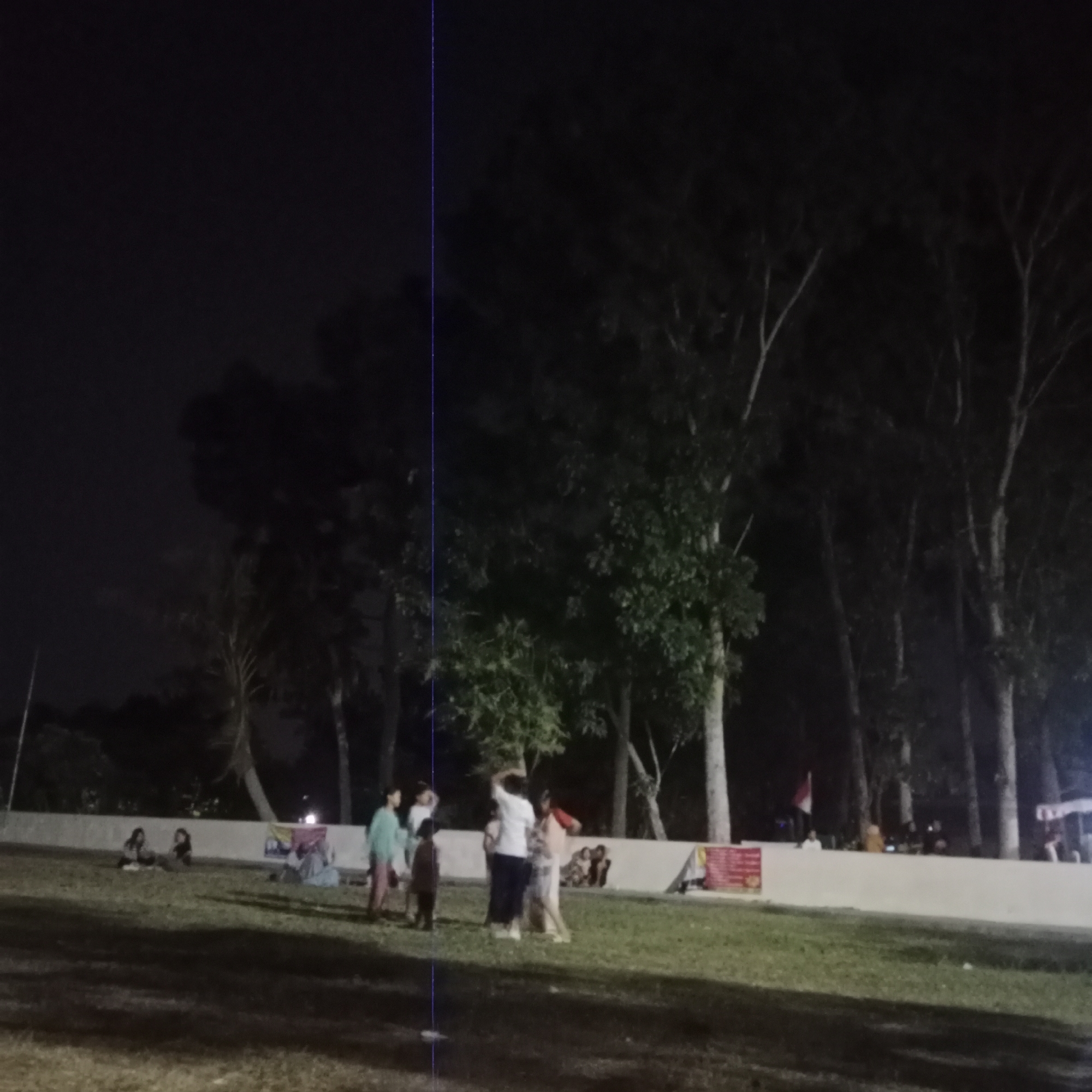 In Kunjeng, Indonesia, at night, a group of people are gathered in an outdoor grassy area near a line of trees. The lighting is dim, suggesting artificial illumination from an unseen source. Several individuals are visible in the foreground and midground, appearing to be socializing or engaging in activities. Some are standing, while others are seated on the grass. A low, light-colored wall runs horizontally across the midground, separating the grassy area from a row of trees behind it. A small Indonesian flag is visible on the wall, and some banners or signs are also present. The overall scene depicts a casual evening gathering in a park-like setting.