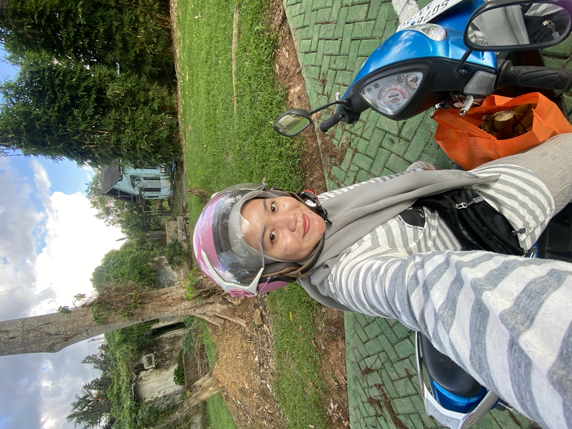 An Indonesian woman wearing a grey hijab and a pink helmet is taking a selfie while sitting on a blue and silver scooter. She is wearing a grey and white striped long-sleeved shirt and a grey scarf. A black fanny pack is visible on her waist. An orange bag containing what appears to be food is hanging from the scooter's handlebars. The scooter is parked on a green tiled path. In the background, there is a large tree, an overgrown abandoned building, and a blue house surrounded by bamboo trees. The scene suggests a residential area on the outskirts of Banjarbaru, Indonesia.