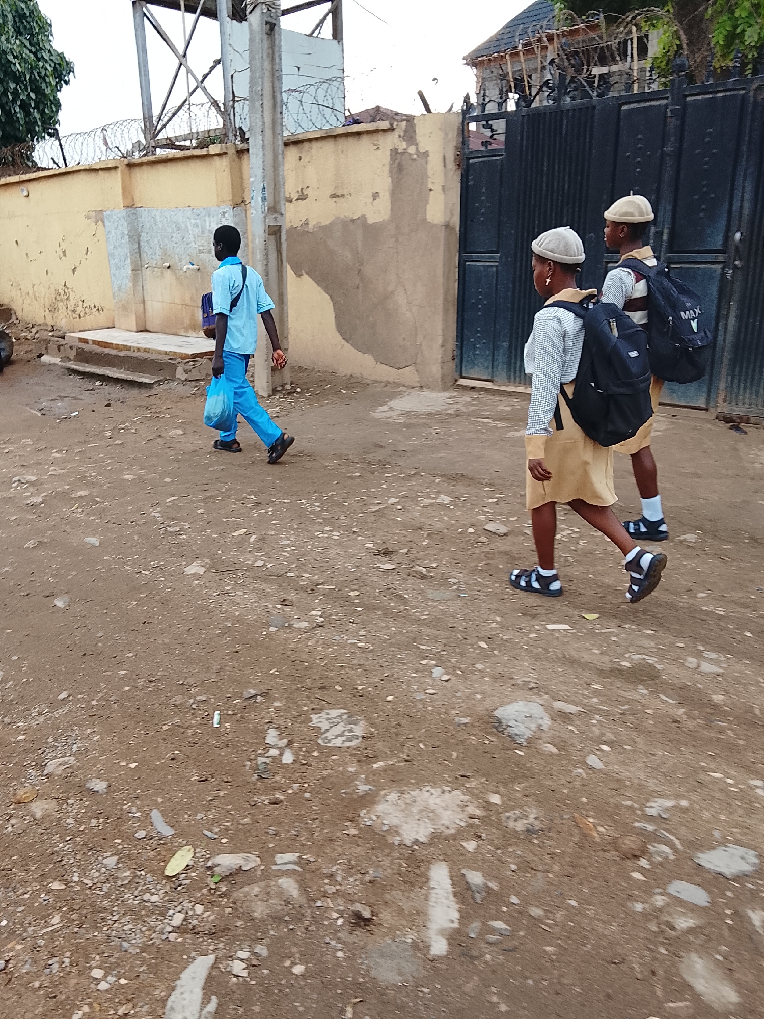 A medium shot, taken outdoors, shows a street in the city of Abuja, Nigeria. The street is unpaved and dirt-covered, with scattered stones and debris. On the left side of the frame, a young man wearing a blue short-sleeved shirt and blue pants walks to the left, carrying a blue plastic bag. On the right side of the frame, two students, dressed in school uniforms, are walking away from the camera. The uniforms consist of khaki skirts, matching hats, white socks, and black sandals. Both students are wearing black backpacks. In the background, a low, yellow building with a barbed wire fence stretches horizontally across the upper portion of the frame. A dark metal gate is visible on the right. A utility pole stands near the building. The lighting appears to be soft and natural.