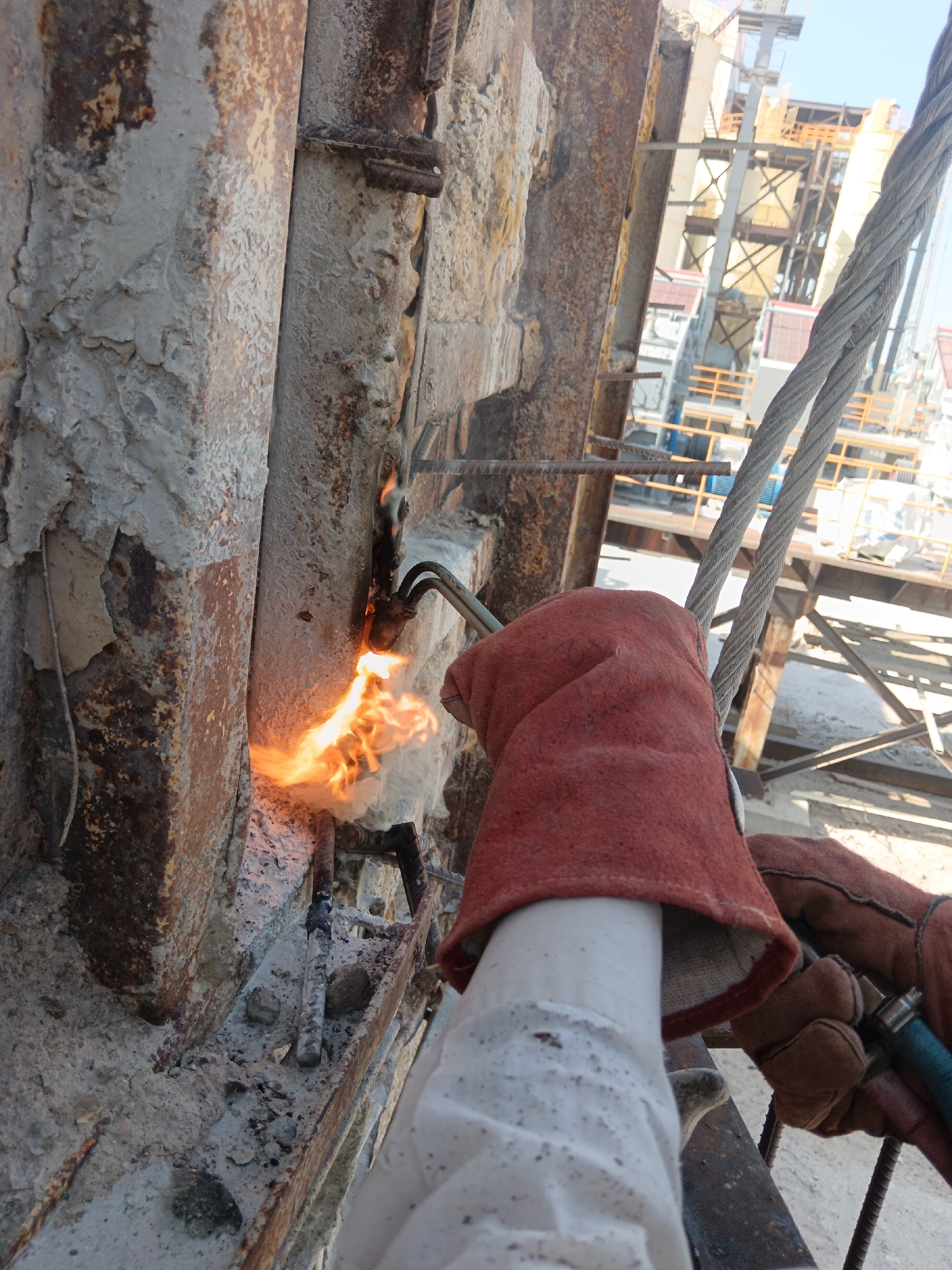 An individual wearing protective gear, including a white undershirt and red welding gloves, is using a cutting torch on a rusted metal structure. The torch flame is bright orange and illuminates the immediate area with intense light and heat. Debris, likely from the cutting process, is scattered around the base of the structure. In the background, a large industrial facility with cranes and scaffolding is visible, suggesting construction or demolition work. A thick cable is also present in the foreground, running diagonally across the image. The scene appears to be outdoors, under a clear sky. The location of Aragatsavan, Armenia is not determinable from the visual information in the image.
