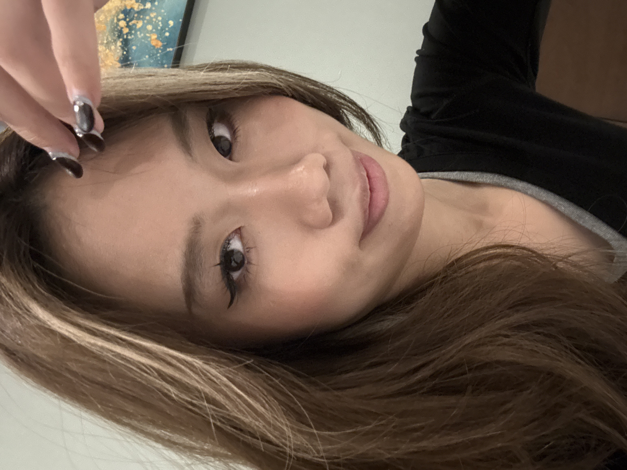 A close-up shot of a woman with long, light brown hair. She is facing the camera at an angle. Her face is in focus, revealing her eyes, nose, and mouth. She has dark eyelashes and pink lips. Her hand is partially visible in the top left corner, with dark fingernails adorned with silver accents. She wears a black top with a grey neckline. A blurry painting of gold and blue hues hangs in the background. There is no evidence to suggest the location is Lanzhou, China.