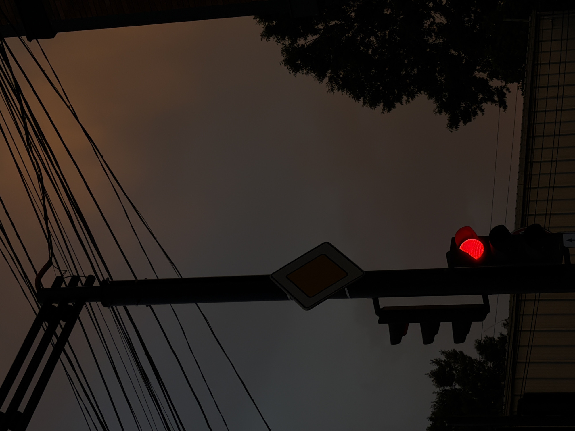 The image shows a street scene at dusk or night in Tbilisi, Georgia. The foreground is dominated by a dark, silhouetted traffic light with a red light illuminated. Below the traffic light, there are three dark, unlit signals, suggesting it's a standard traffic signal. The traffic light is mounted on a horizontal bar which is supported by a dark structure with many electrical wires fanning out to the left. A diamond-shaped road sign with a yellow center and white border is visible on the same horizontal bar, to the left of the traffic light.
In the background, the sky is a muted, hazy orange-brown, indicating a low light condition, possibly due to pollution or an overcast sky. Silhouetted against the sky on the right side of the image is the edge of a building with a grid-like pattern, possibly a fence or a window grate. Dark tree foliage is visible in the upper right and lower right corners of the frame, also silhouetted. The overall mood is somewhat somber and urban, with the red traffic light being the only source of vibrant color. There is no visible text in the image.