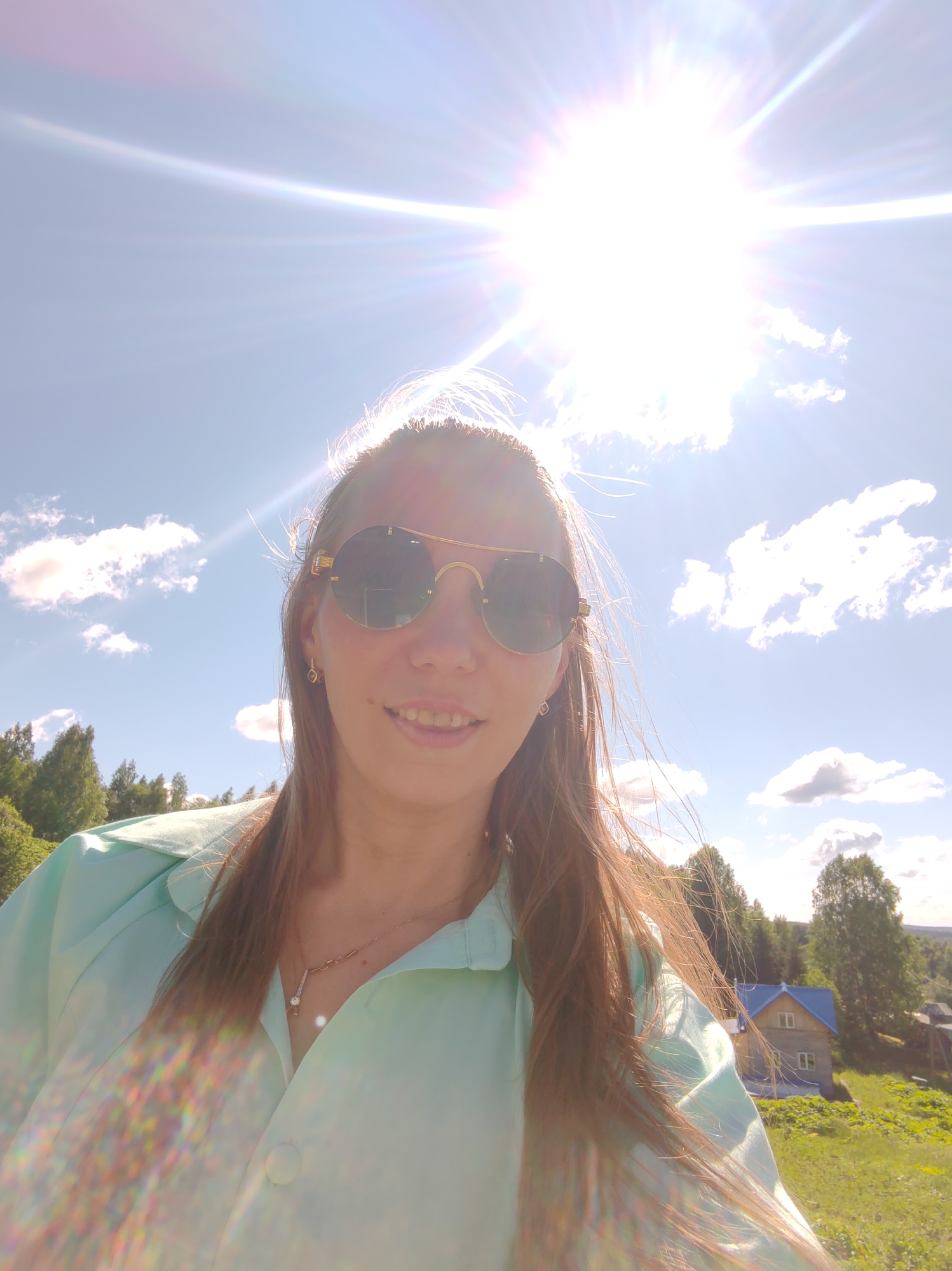 A young woman with long brown hair and sunglasses is captured in a selfie outdoors under a bright, sunny sky with scattered clouds. She is wearing a light blue collared shirt and a necklace with a pendant. In the background, a wooden house with a blue roof is visible, nestled amongst green trees and rolling hills. The scene suggests a rural or suburban setting, potentially in the city of Chukhlem, Russia, given the architectural style of the house and the natural landscape. The sun is intensely bright, creating lens flare and washing out some details in the upper portion of the image.