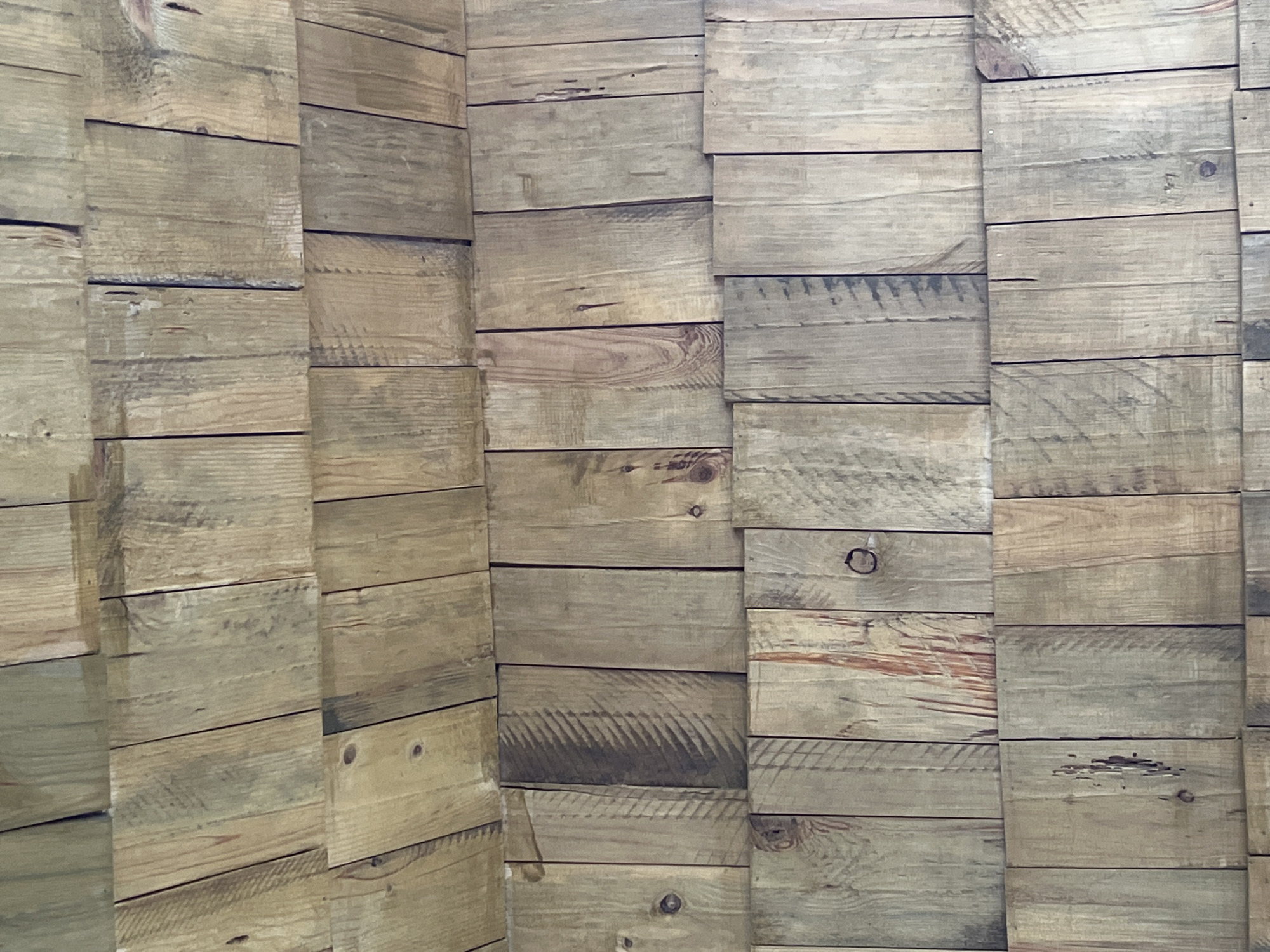 The image displays a close-up, full-frame view of a wall constructed from weathered, vertically-oriented wooden planks. The planks vary in width and color, showing a natural, rustic aesthetic with visible wood grain, knots, and occasional imperfections such as small holes or darker discoloration. The arrangement suggests a shingle-like pattern where the upper planks overlap the lower ones, creating horizontal lines of separation. The lighting is diffuse, highlighting the texture and natural variations in the wood. There are no people or other discernible objects in the image. The scene is of an exterior or interior wall, likely in Daulėnai, Lithuania.