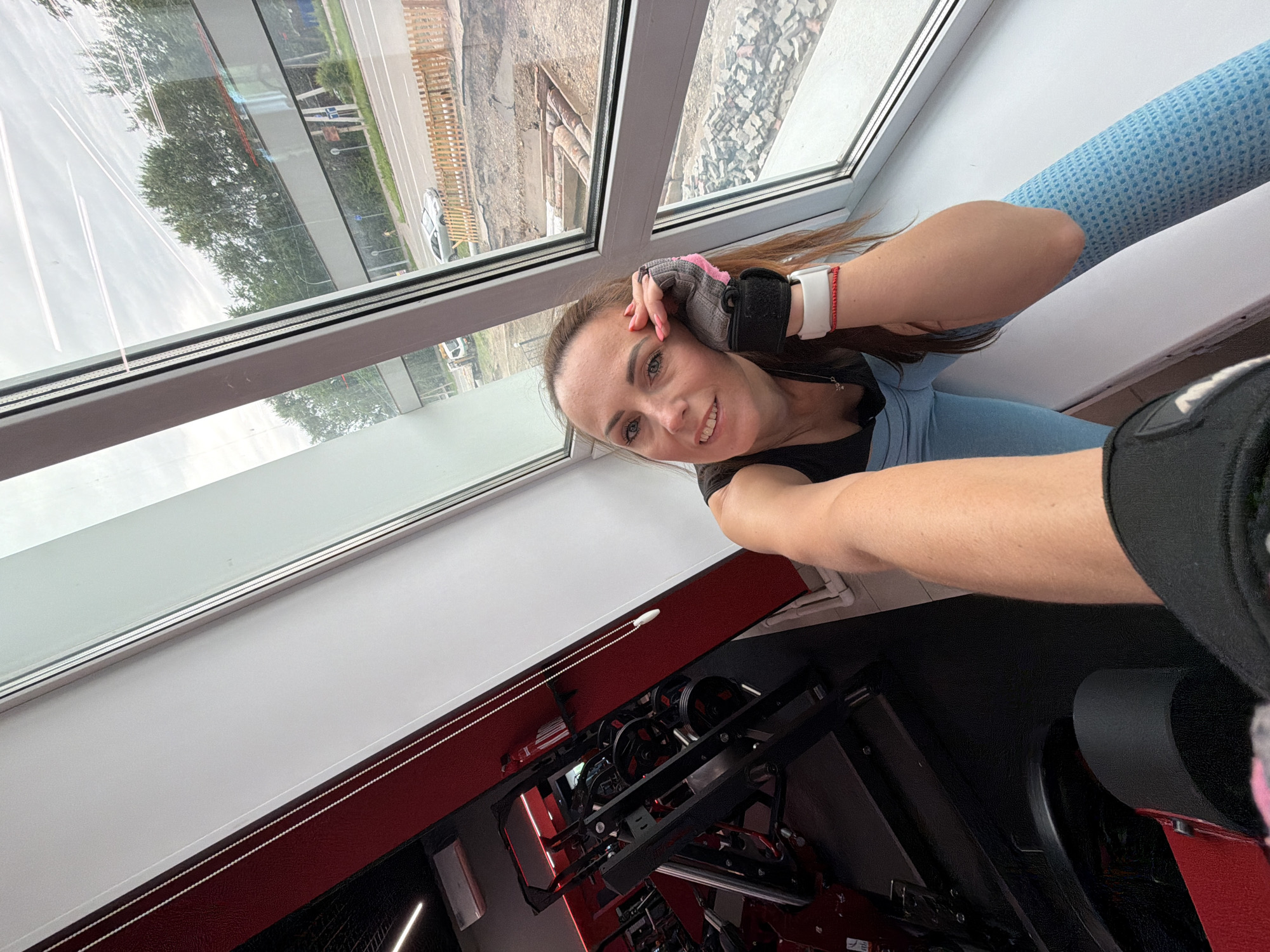 In Birobidzhan, Russia, a woman with auburn hair and visible makeup is posing for a selfie in a gym. She is wearing a black tank top, blue leggings, and pink and grey workout gloves. Her right arm is resting on her forehead, and her left arm is extended towards the camera. The background shows gym equipment, including a weight machine, and a large window overlooking a street with trees, cars, and construction. The lighting is bright, suggesting it is daytime.