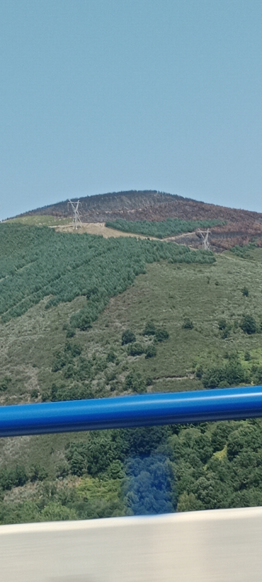 The image shows a hillside in Vega de Valcarce, Spain. The hillside is covered with trees, with some areas appearing burnt or dry. Two tall, metal pylons with power lines are visible on the hillside. The sky is clear and blue. A blue railing is in the foreground, suggesting the photo was taken from a moving vehicle or elevated viewpoint.