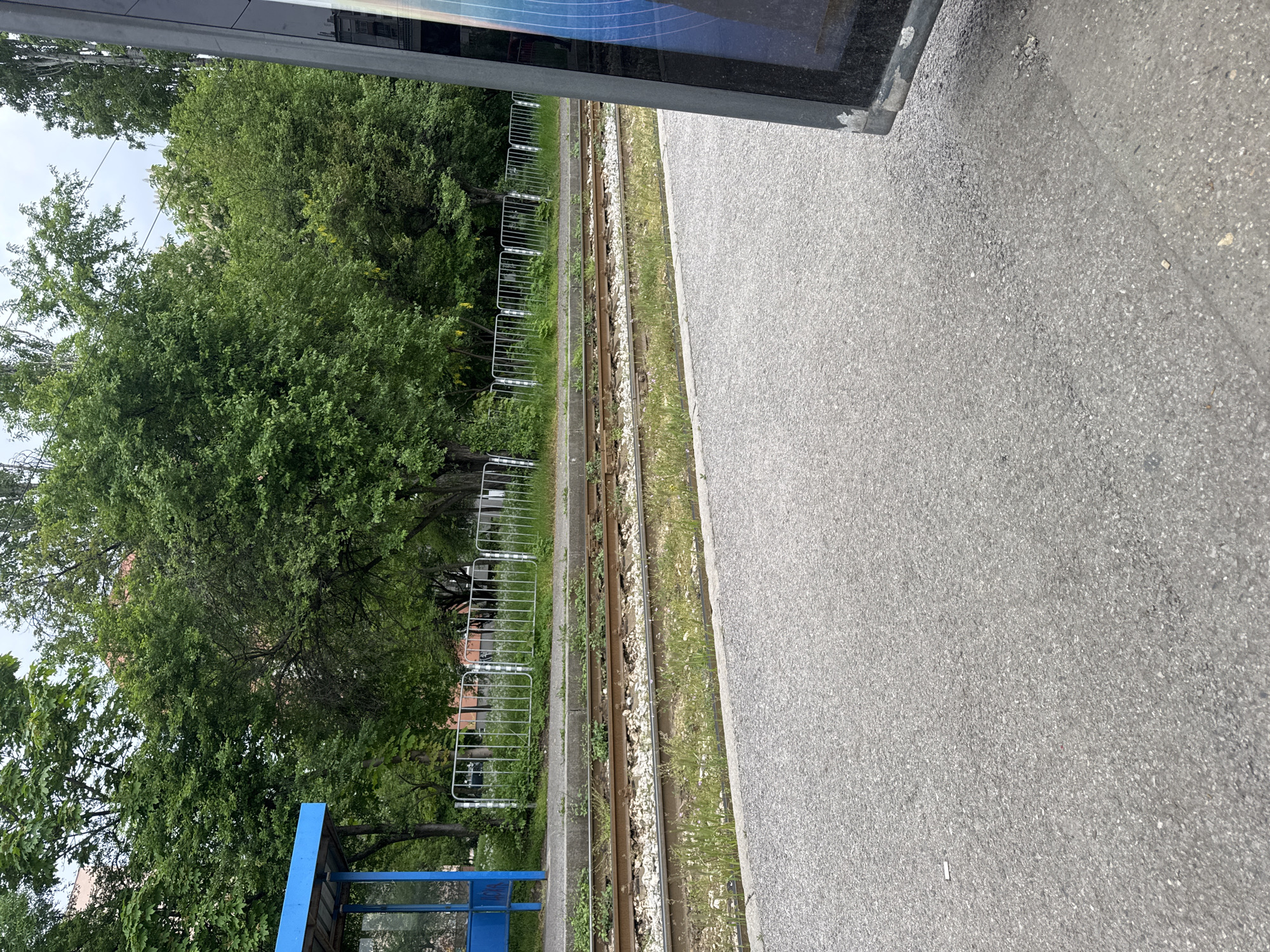 The image shows a railway track alongside a paved area, likely a platform. The track is lined with small white stones and some vegetation. On the other side of the track, there is a silver fence and dense green trees. In the foreground on the left, a blue bus stop shelter is visible. The location is in the city of Unknown, Unknown. The image appears to be taken from a slightly elevated position, possibly from inside a vehicle or building.