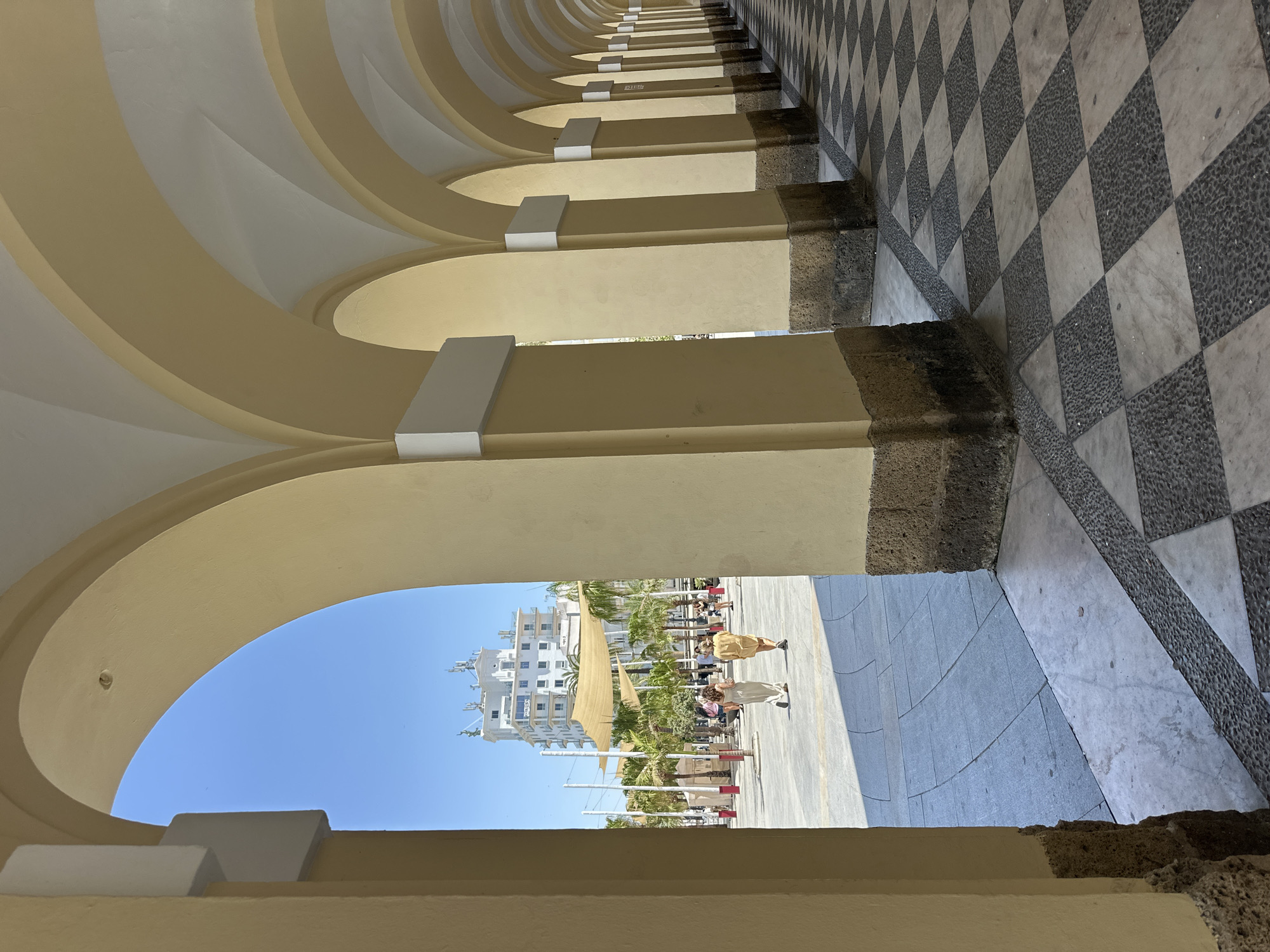 The image depicts a view through an arched portico with pale yellow columns and a checkered floor pattern of dark gray and white tiles. The portico extends into the distance, creating a sense of depth. Through the arches, a bright outdoor plaza is visible under a clear blue sky. Several palm trees are scattered across the plaza, along with people walking and standing. A prominent white building with multiple stories and balconies stands in the background, topped with antennas. Two women are walking away from the camera, towards the plaza. The architectural style and the presence of palm trees suggest a Mediterranean or southern European location, consistent with Cádiz, Spain. The lighting indicates it is daytime, likely with strong sunlight.