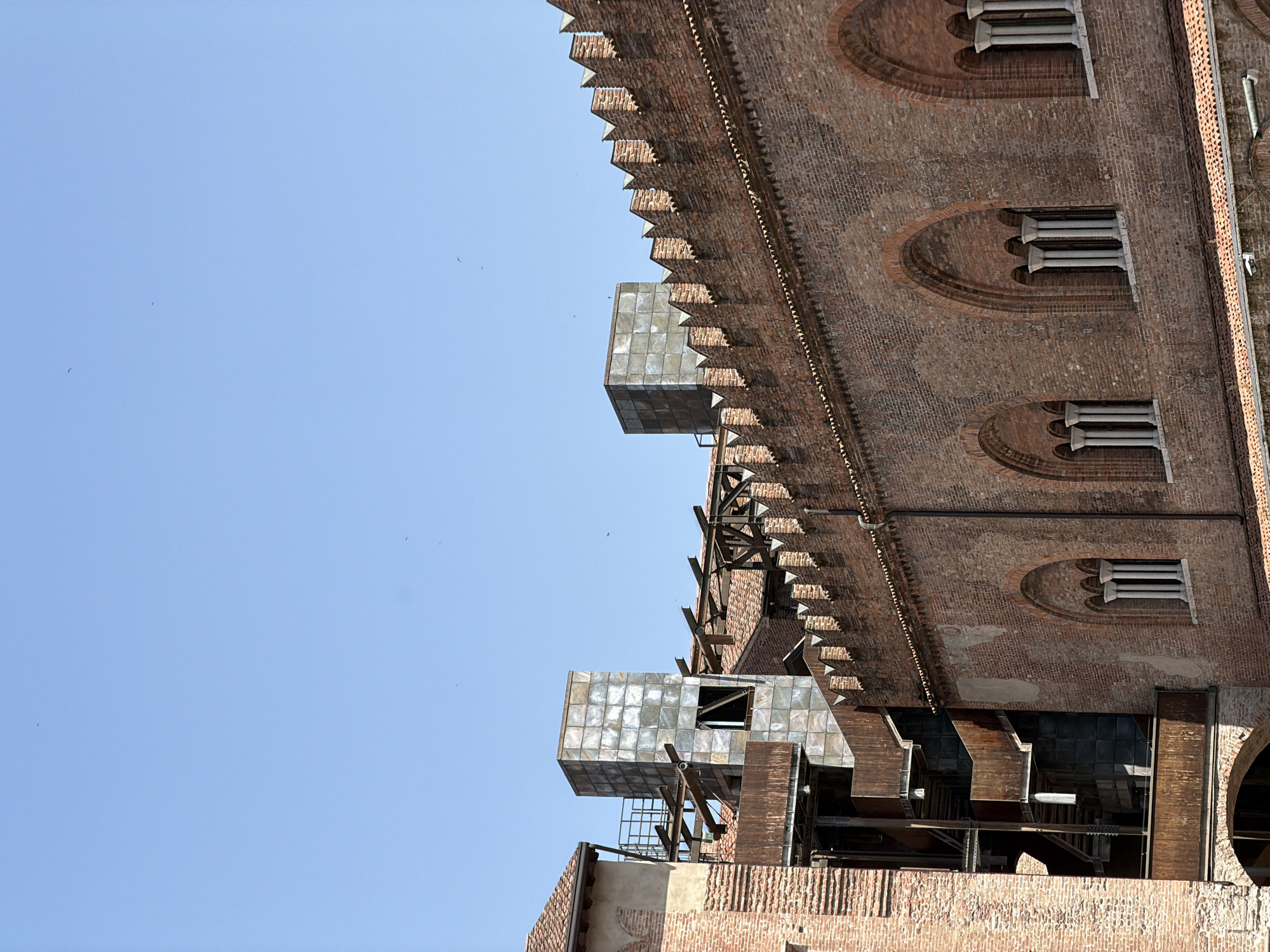 A worm's eye view, taken in Mantua, Italy, showcases the facade of a brick structure, possibly a historic building or tower. The building is adorned with a crenellated edge, with metal-clad, cube-like additions protruding from the brickwork. Windows, defined by arched frames and vertical columns, punctuate the facade. Metal beams and scaffolding are juxtaposed against the brick exterior. The background is a clear, blue sky with a few small birds visible.