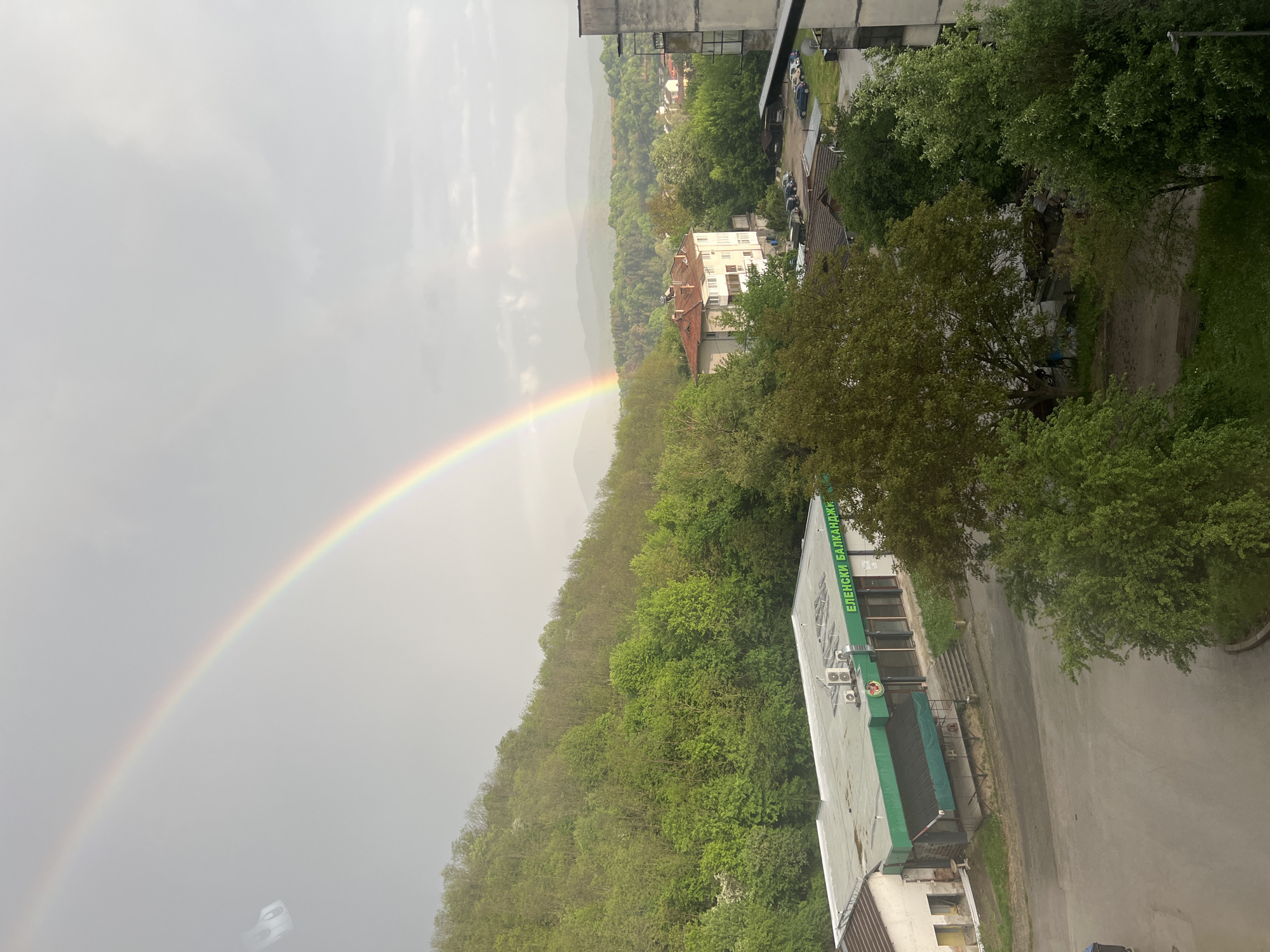 The image depicts a view from an elevated position in Elena, Bulgaria, featuring a vivid rainbow spanning across a cloudy sky. The foreground showcases the urban landscape, with buildings partially visible on the right, and lush green trees covering the hillside. One building has a sign written in Bulgarian that reads “ЕЛЕНСКИ БАЛКАНДЖИ,” suggesting a local business. In the distance, mountains and a small town are visible under the cloudy sky. The street adjacent to the building appears wet, indicating recent rain, which is likely the cause of the rainbow. The overall scene is a blend of urban and natural elements under atmospheric conditions.