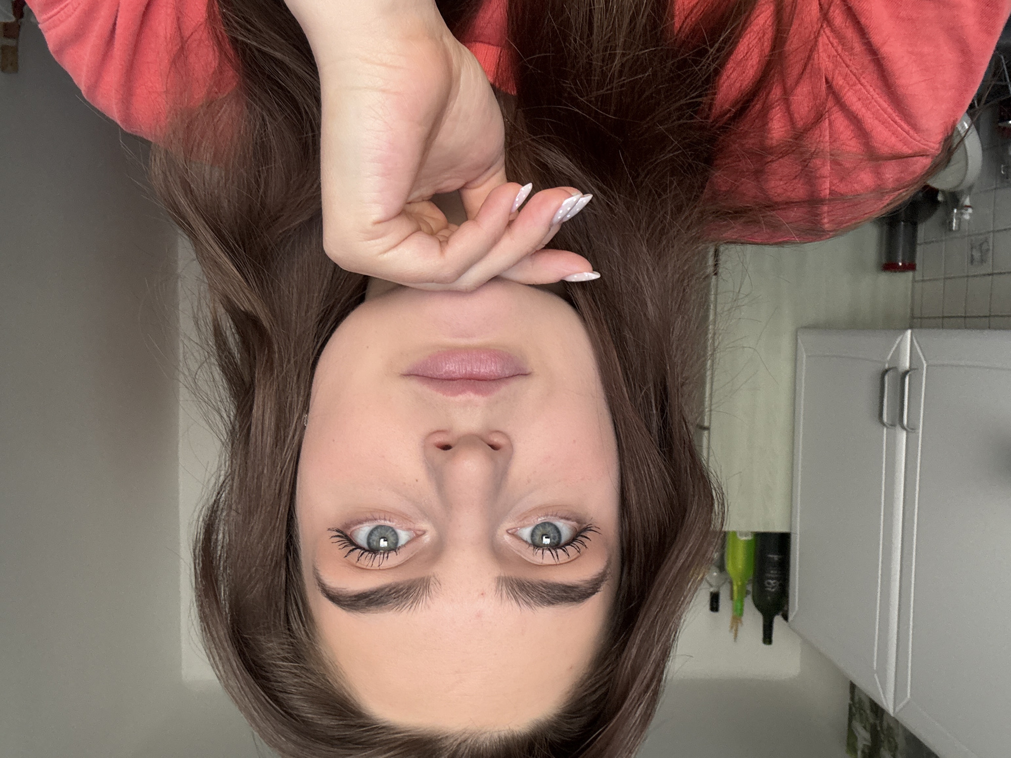 An upside-down portrait features a young person, likely female, with fair skin, long dark brown hair, and prominent light blue-grey eyes. She appears to be lying down, looking directly into the camera with a neutral and calm expression. Her lips are slightly parted and have a natural pink color. Her eyelashes are accentuated with mascara, and her eyebrows are dark and well-defined.
Her right hand is gently positioned over her forehead, partially obscuring her hair. Her nails are long, almond-shaped, and painted white with small, light-colored polka dots. She is wearing a coral-red, textured long-sleeved top or sweatshirt, which frames her face.
The background, also inverted, suggests an indoor home environment. To her right (left in the image), a plain light grey wall is visible. To her left (right in the image), elements of what appears to be a kitchen or utility area are present, including white cabinet doors with silver bar handles, a light-colored tiled backsplash, and a red and black handheld vacuum cleaner mounted on the wall. Partially obscured by her hair are glimpses of bottles, one distinctly green, suggesting items on a counter or shelf.
The lighting is soft and even, suggesting either natural daylight or well-diffused artificial light, creating a clear and well-lit scene. No discernible text is visible in the image.