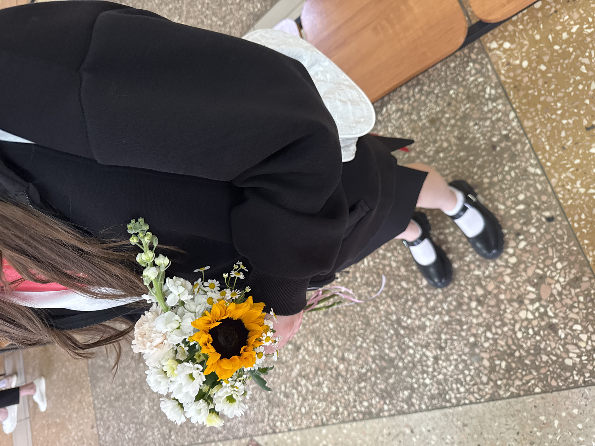 A person is seen from an overhead angle, seated, and holding a bouquet of flowers. The bouquet features a sunflower, white carnations, and daisies. The person is wearing a black jacket and dress with white socks and black Mary Jane shoes. A white purse is partially visible near the person's shoulder. The person has long brown hair. The floor has a terrazzo tile pattern. The location is Ivanovo, Russia.
