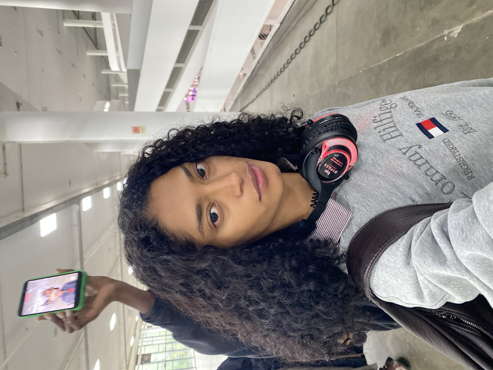 A woman with curly dark hair is shown in a vertical, tilted selfie. She is light-skinned and gazes directly into the camera with a calm expression. She is wearing a gray Tommy Hilfiger sweatshirt and has black and red headphones around her neck. A dark leather jacket is draped over her shoulder. In her outstretched left hand, she holds a smartphone with a green case, displaying a picture of someone on the screen. The setting appears to be an indoor public space with a high ceiling, white walls, and linear lighting fixtures. A chain link fence runs along the upper right of the frame. The photo was possibly taken in the city of São Paulo, Brazil.