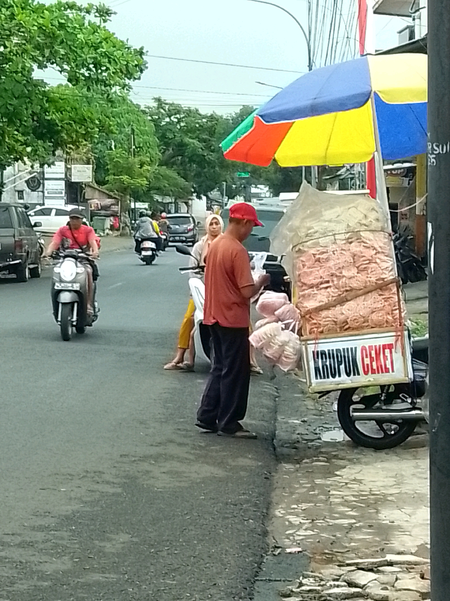 The image depicts a street scene in Jember, Indonesia, likely a roadside where vendors operate. In the foreground, a man wearing a red cap and an orange t-shirt stands next to a motorcycle cart laden with packaged snacks, identified by a sign that reads "KRUPUK CEKET". The cart is covered by a multi-colored umbrella. The man appears to be attending to his wares. Behind him, other people are visible, including a person riding a motorcycle and another on foot. The environment shows a paved road, some greenery in the background, and buildings along the street. The overall impression is of a typical urban commercial area with mobile vendors.