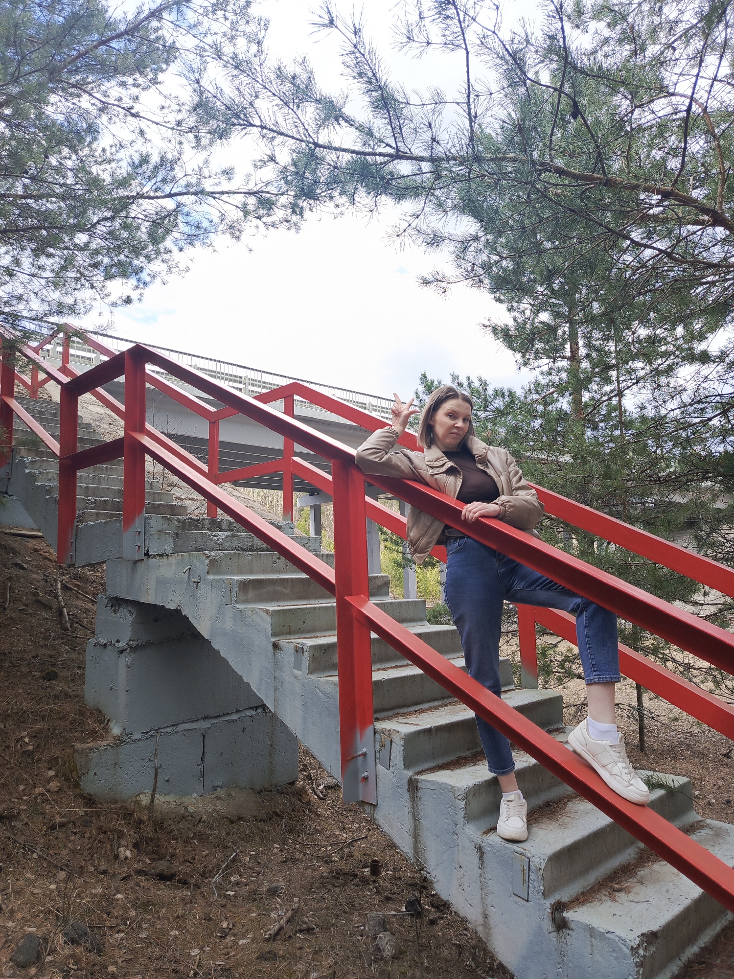 The image features a young woman posing playfully on a set of grey concrete stairs with striking red metal railings, set outdoors. She has fair skin, light brown hair, and is dressed in a light brown or beige jacket over a dark top, blue jeans, white socks, and white sneakers. With a casual pose, she leans on the red railing with her right elbow, her left leg bent and foot resting on the railing. She forms a "peace" sign with her left hand and sports a playful pout or grimace.
The stairs, appearing somewhat weathered, ascend towards a bridge or overpass structure visible in the background, suggesting a connection to infrastructure. The vibrant red railings contrast sharply with the grey concrete and the surrounding natural elements. Pine trees with their characteristic needles and branches frame the top of the image and form the backdrop for the woman, indicating a wooded or forested area. The ground at the base of the stairs is covered with dry pine needles, leaves, and earth. The sky above is bright and appears overcast or partly cloudy, providing soft, diffuse lighting characteristic of daytime. No visible text is present. The setting is consistent with a natural environment found in Chaikovsky, Russia.