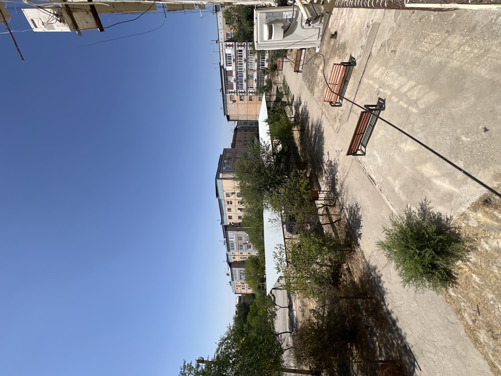 The image depicts a sunny day in a courtyard area in the city of Şamaldı-Say, Kyrgyzstan. In the foreground, a concrete patio area is visible, with two parallel wooden benches with metal frames situated on it. A single green bush is planted in a patch of dry grass to the left of the benches. In the midground, a covered parking or seating area with a light-colored metal roof is present. Beyond this, a row of apartment buildings of varying heights and architectural styles stretches across the horizon under a clear blue sky. These buildings are several stories tall and appear to be made of brick and concrete. Green trees and bushes are scattered throughout the courtyard and surrounding areas, casting shadows on the ground. An outdoor air conditioning unit is mounted on the side of a building on the right edge of the frame. The overall scene conveys a sense of a residential urban environment on a warm, clear day.