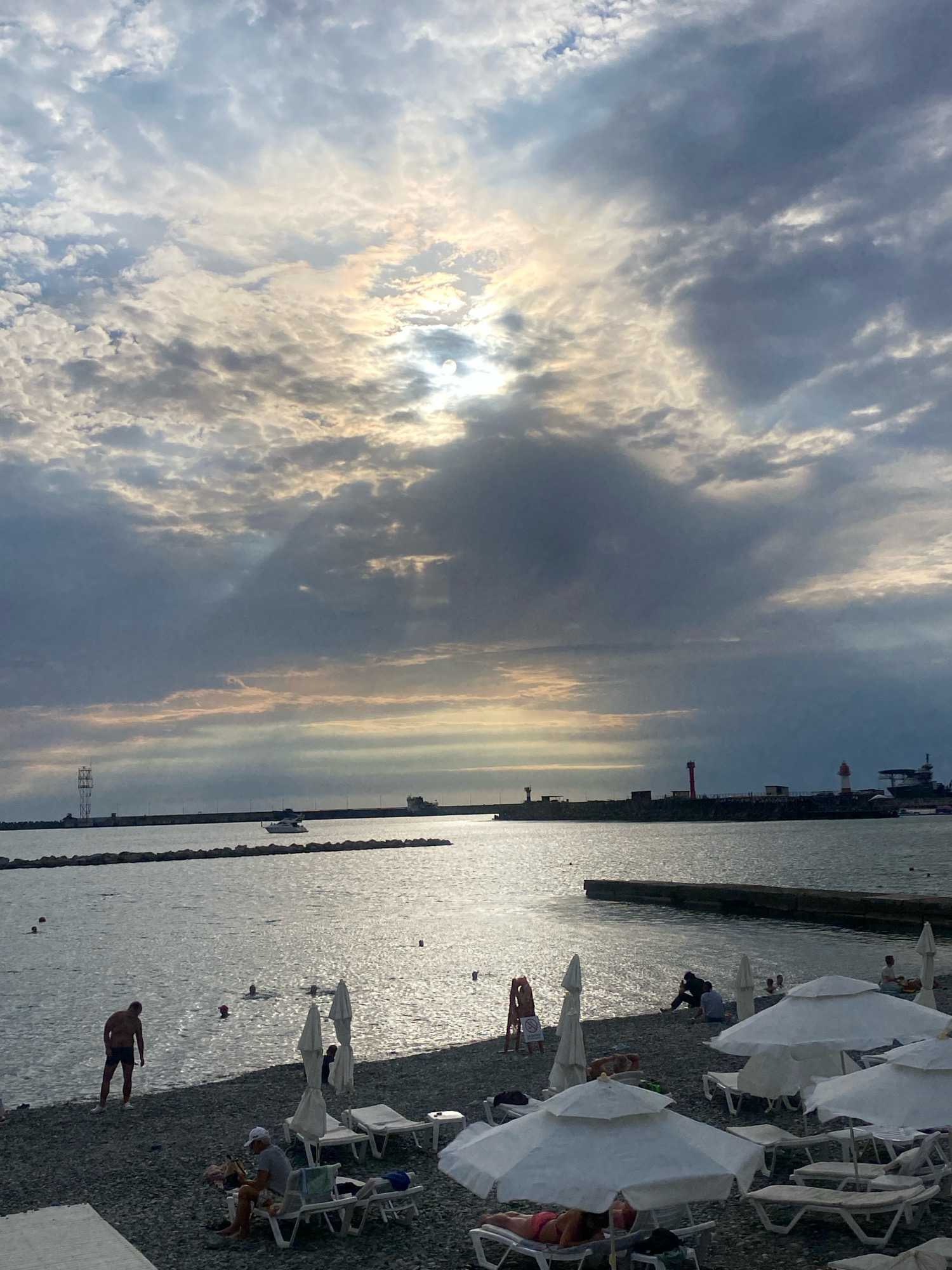 The image depicts a view of a pebbled beach and the sea in Sochi, Russia, under a dramatic sky filled with clouds. The sun is partially visible through the clouds, casting rays of light onto the water. Several people are present on the beach, some relaxing on lounge chairs under white umbrellas, while others are in the water swimming or wading.
In the foreground, lounge chairs and umbrellas are arranged on the pebble beach. A man is standing near the water's edge, facing away from the camera. Further out, several people are visible in the sea, appearing to be swimming or enjoying the water.
The sea stretches towards the horizon, where a breakwater extends into the water. A small white boat is sailing on the water near the breakwater. On the breakwater, there are structures, including what appears to be a telecommunication tower on the left and two lighthouses on the right, one red and white, and another smaller white one. The overall impression is of a somewhat overcast but still active day at a coastal resort.
