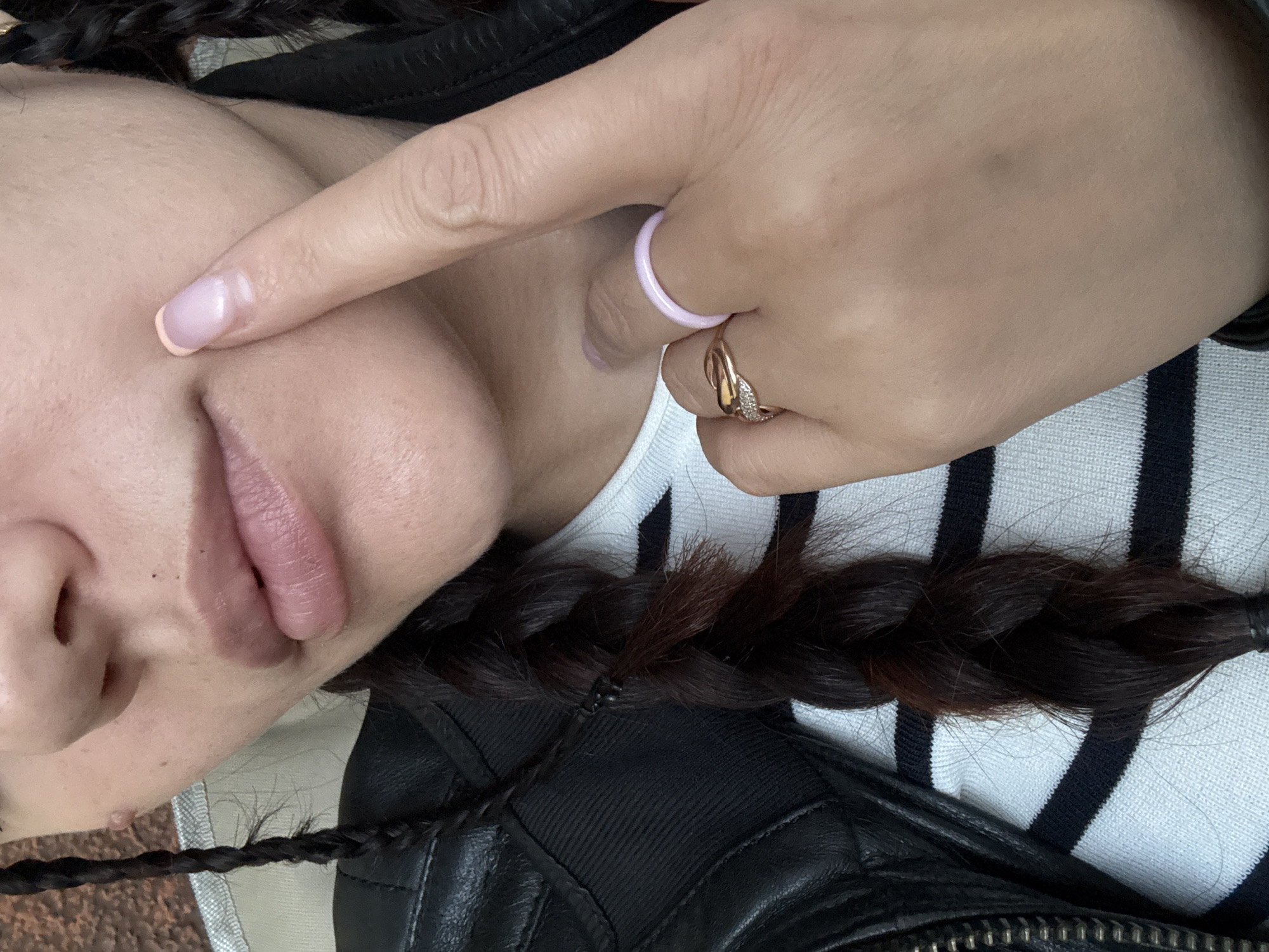 A close-up, medium shot shows a person's mouth, chin, and hand. The person's lips are slightly parted and appear to be a natural pink color. A finger with a light pink manicured nail is touching the person's cheek near the mouth. Two braids of dark brown hair are visible on either side of the person's face, and one braid extends down the chest. The person is wearing a black leather jacket over a white and navy striped shirt. Two rings are on the person's finger; one is a thin, light pink band, and the other is a rose gold band with small diamonds. The background is blurred and appears to be indoors. The scene is static and does not depict any discernible action or interaction.