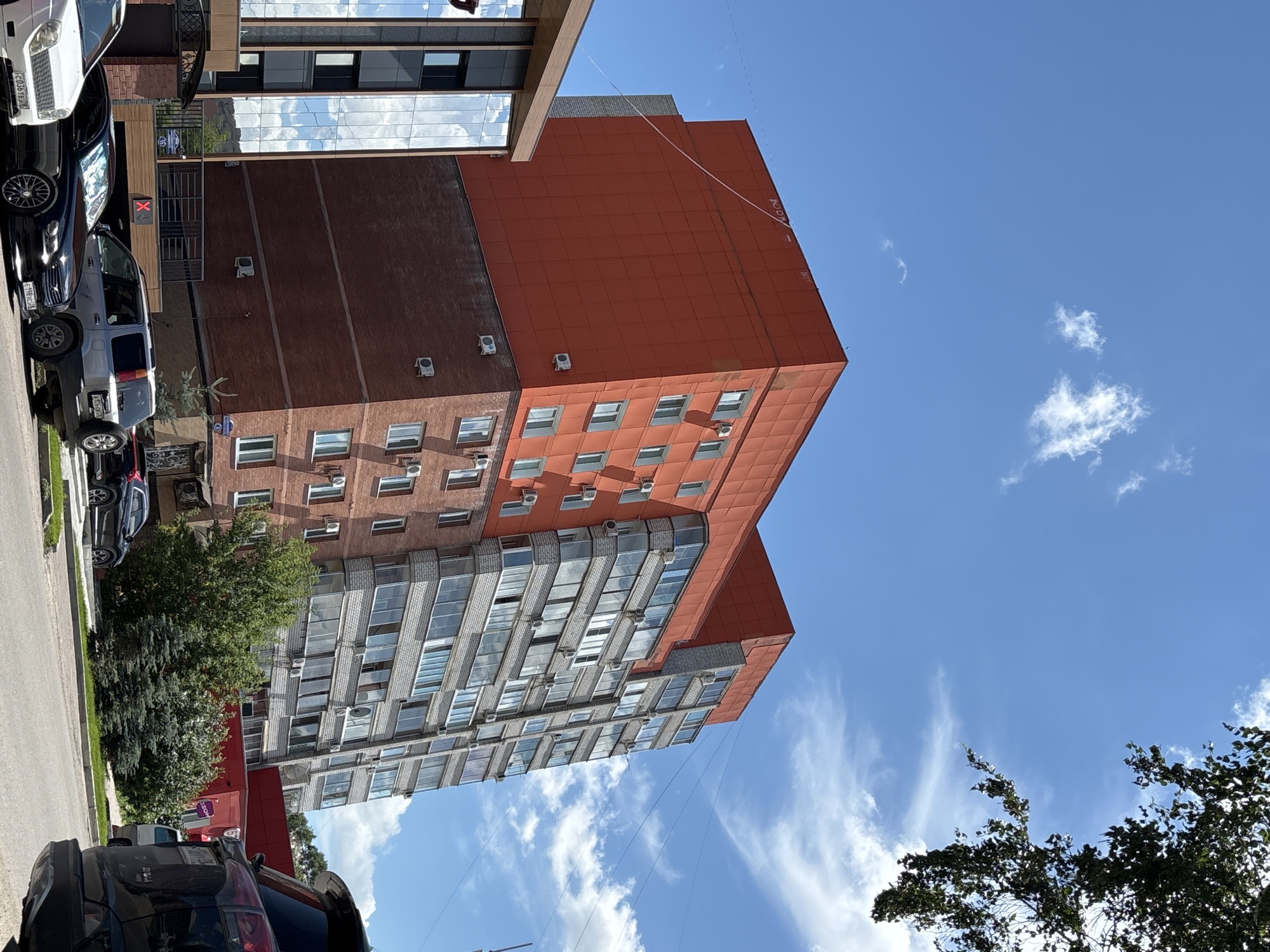 This image depicts a daytime street scene in Blagoveshchensk, Russia. In the background, a tall, modern apartment building dominates the skyline. The building has a distinct design, with the upper floors clad in bright orange panels and the lower floors constructed of red brick. The upper section features numerous windows, many with balconies, and appears to have an angular design. The lower portion of the building is made of red brick and has a more traditional facade with windows and air conditioning units. To the left of the orange building, a contemporary glass-fronted building with dark framing is visible, reflecting the sky and clouds.
In the foreground, several cars are parked along a street. These include a white sedan, a black sedan, a silver SUV with a spare tire mounted on the back, and a black SUV partially visible on the right. Green trees and bushes are interspersed along the street and around the buildings, suggesting a semi-urban or residential area. The sky is a clear blue with scattered white, fluffy clouds. The overall impression is of a sunny day in a Russian city.