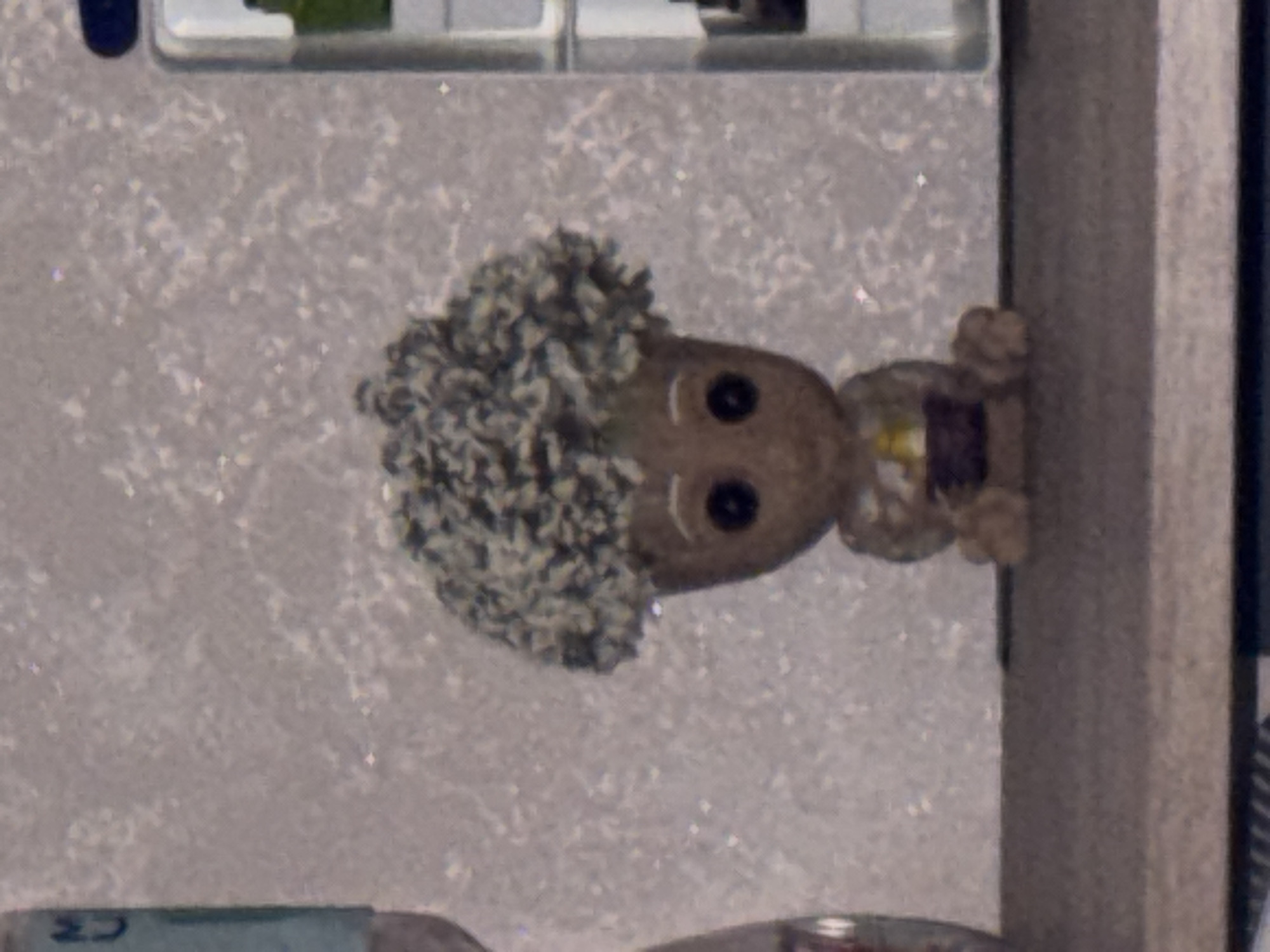 The image depicts a close-up, eye-level, medium shot of a small, seated figurine with a large, bushy, textured head resembling moss or leaves. The figurine has dark, circular eyes and a simple, curved line for a mouth. It appears to be made of a light brown material and is wearing what looks like a brown shirt with a yellow or orange rectangular design on the chest. Its legs are bent, and its arms are resting in its lap. The figurine is positioned on a dark, flat surface, possibly a shelf. The background is textured and appears to be a wall with a speckled or granular pattern in shades of light grey and white. To the left of the figurine, a portion of a light blue cylindrical object with some writing, possibly a bottle or label, is visible. To the right, a white vertical object with a darker outline, suggesting a frame or edge of another item, is present. The overall lighting is soft and diffused, with no harsh shadows. There is no identifiable information indicating the location within the city of Saratov, Russia.