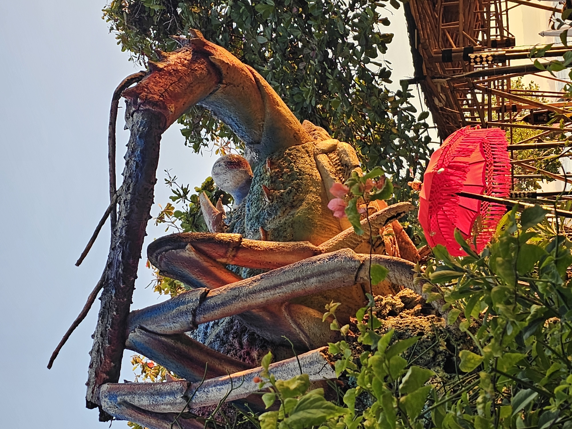 The image shows a large, decorative crab sculpture in Denpasar, Indonesia. The sculpture is primarily brown, blue and green. It is partially obscured by green foliage, including climbing plants and flowering shrubs. A red paper lantern hangs near the crab sculpture. Behind the lantern and sculpture is a bamboo structure. The sky is a light blue-gray color.