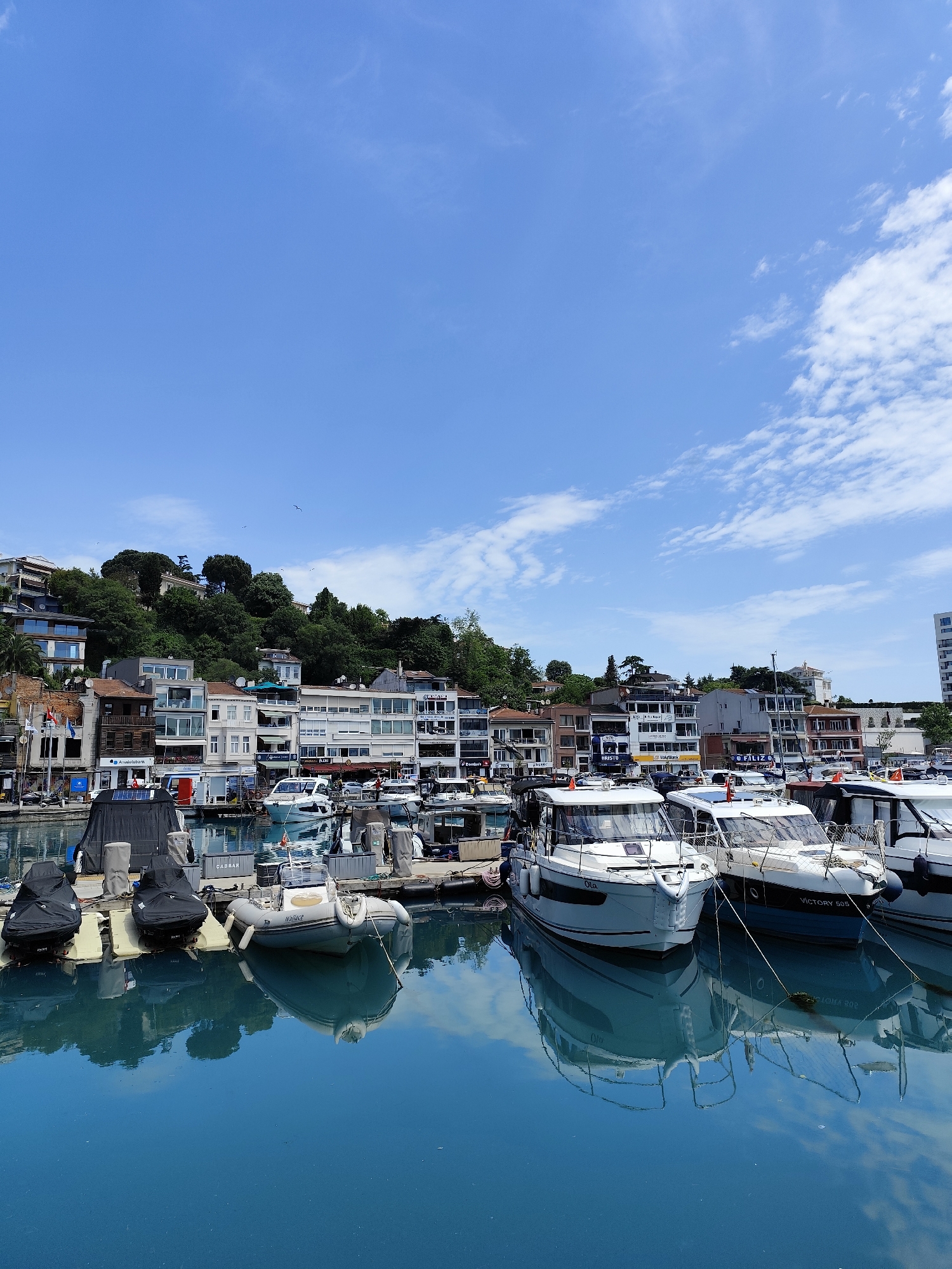 This image captures a vibrant, sunny day at a marina in Sarıyer, Turkey, featuring a collection of boats docked alongside a hilly shoreline adorned with buildings and lush greenery.
The upper portion of the frame is dominated by a clear, bright blue sky with sparse, wispy white clouds, indicating fair weather. Below the horizon, a cluster of buildings with diverse architectural styles, ranging from modern structures with large windows to more traditional, multi-story houses, are nestled into a verdant hillside. The hillside is densely covered with trees and foliage, appearing vibrant green.
In the foreground and midground, numerous boats are moored in the calm, turquoise water. The water acts as a mirror, perfectly reflecting the boats, the blue sky, and hints of the shoreline architecture. The boats are primarily modern white motor yachts and cruisers of varying sizes, interspersed with smaller inflatable dinghies, some covered with black tarps. Many boats feature railings and small cabins.
No people are explicitly visible, suggesting a peaceful, uncrowded scene. The overall atmosphere is tranquil and picturesque, likely capturing a leisurely moment during daylight hours, given the bright sun and clear visibility.
Visible text includes "Anadolubank" on a building sign on the left side of the shore. On one of the white boats towards the right, the name "VICTORY 505" is clearly readable on its hull, while another boat appears to have "Ota" written on its bow. A container on a dock between boats displays "CABBAR".