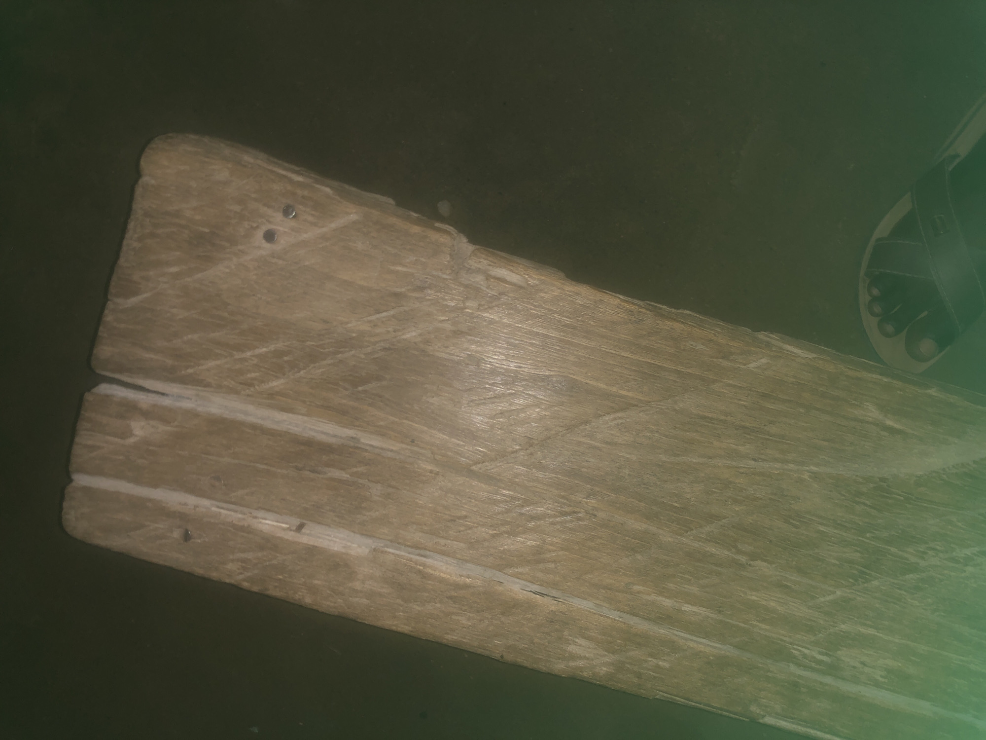The image shows a close-up, angled view of two weathered wooden planks joined together. The wood is light brown with visible grain and some scratches and imperfections. There are visible nail heads on the surface of the planks, one on the left plank and two on the right plank. The background is a dark, out-of-focus area that appears to be a floor or ground. In the lower right corner, a portion of a person's foot wearing a black sandal is visible. The scene appears to be indoors or in a shaded outdoor area, given the lighting. There is no specific information within the image to indicate its location in Jalingo, Nigeria.