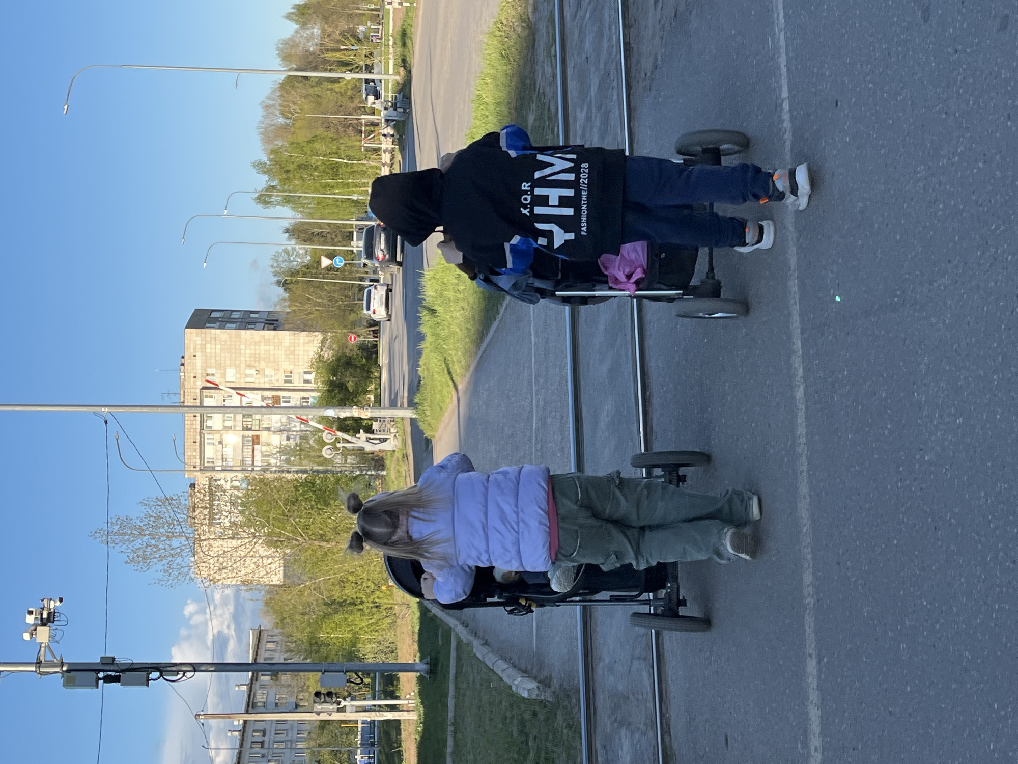 Two children are pushing baby carriages on a paved surface, likely a road or sidewalk, with a tram track running along it. One child is wearing a black hooded sweatshirt with the text "X.Q.R FASHIONTHE//2028" on the back and blue pants. The other child has blonde hair in two pigtails and is wearing a lavender jacket and green pants. The environment includes apartment buildings, streetlights, and a level crossing signal, indicating a residential area near a railway or tram line. The presence of level crossing suggests that it is Chaikovsky, Russia.