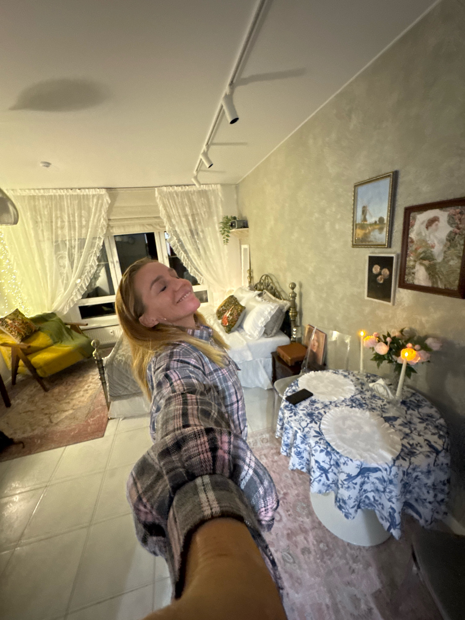 The image depicts a bedroom setting, with a woman in the foreground taking a selfie. The room is decorated in a rustic and slightly vintage style. A bed with white linens and decorative pillows is visible in the background, next to a nightstand. A round table covered with a blue and white patterned tablecloth is set with two white placemats and a vase of pink flowers with lit candles. On the wall, there are several framed pictures. A yellow armchair with decorative pillows is placed near a window covered by white lace curtains. The floor is tiled. The overall impression is that of a cozy, personalized space.