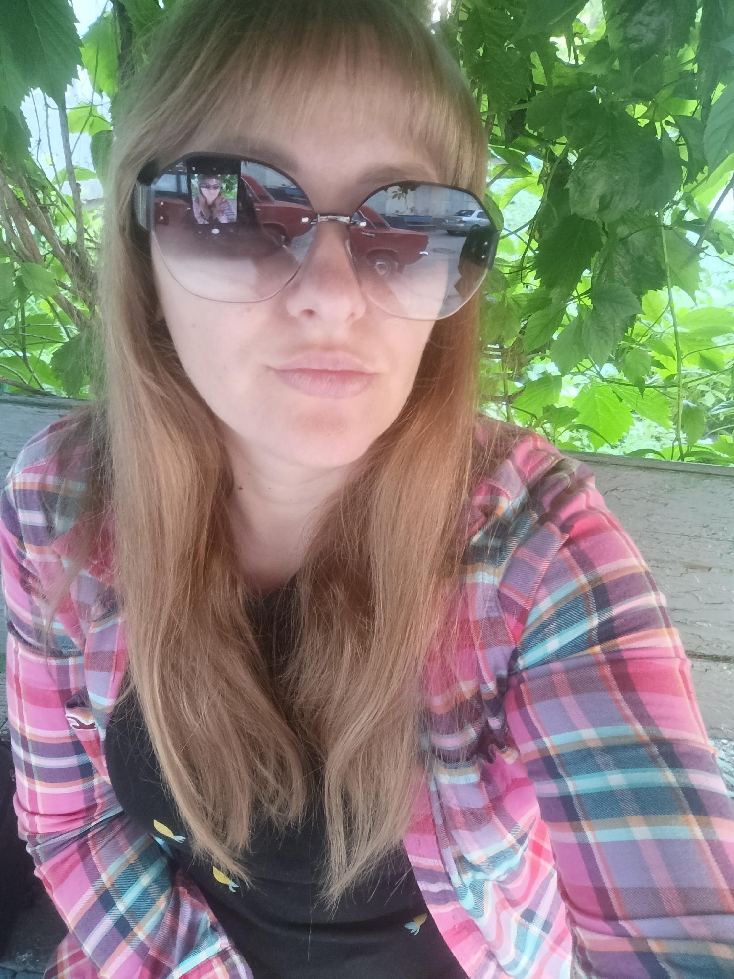 A close-up, selfie-style shot captures a person wearing large, round, gradient sunglasses and a plaid shirt. The person has long, blonde hair and is looking directly at the camera. Their lips are slightly parted. The sunglasses reflect a scene of a street with cars, and the person's own face. Behind the person, lush green foliage is visible against a light-colored, textured background that appears to be a wall or wooden structure. The image is taken outdoors, with natural light.