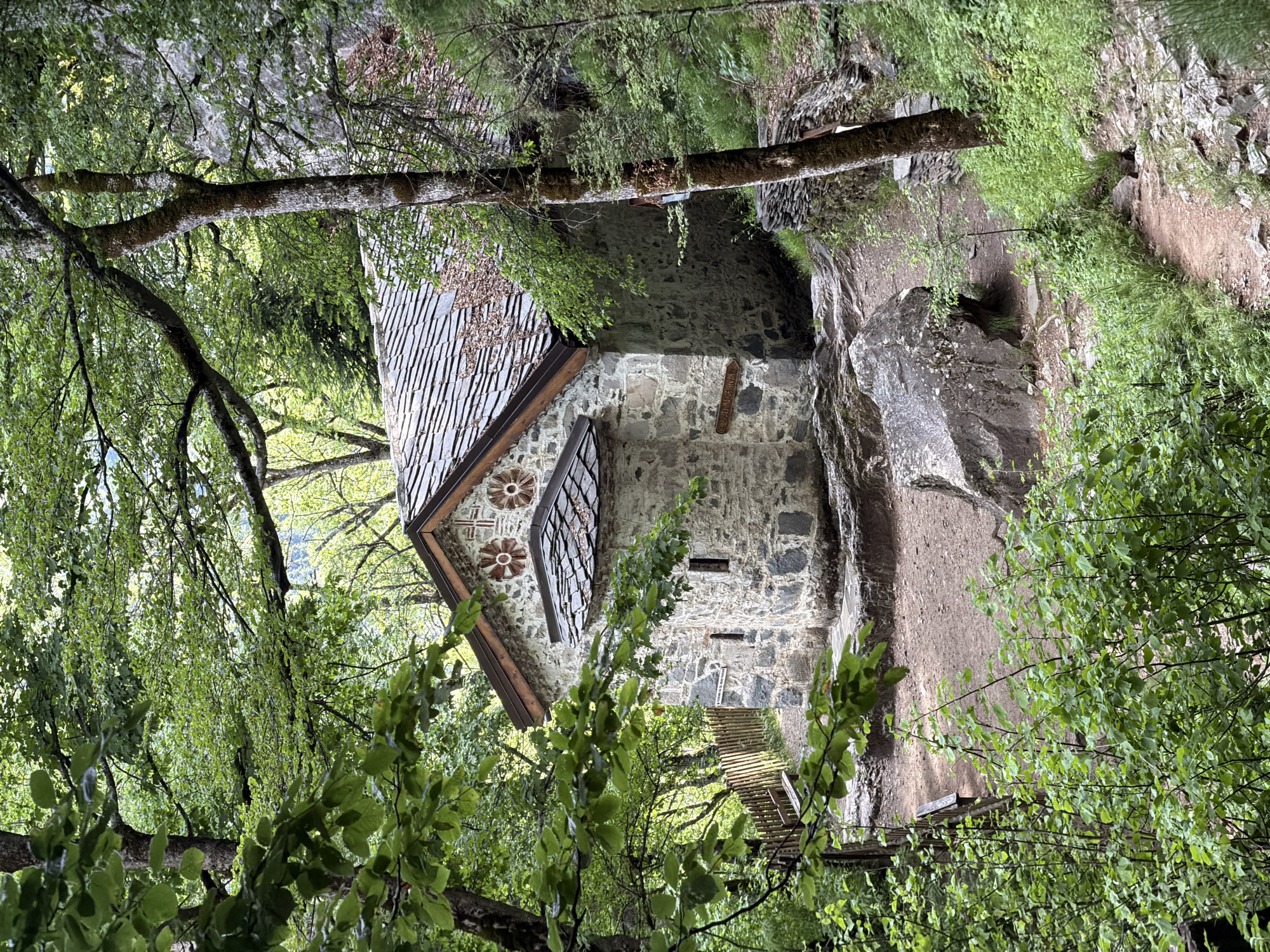 A stone building with a gray tiled roof is nestled in a forest, possibly near a rock face. The building has decorative floral designs on its front façade. A wooden walkway appears to lead to the building. The image may be of the St. George the Victorious Church in Govedartsi, Bulgaria.
